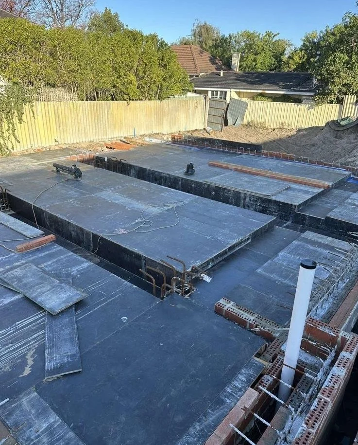 Construction site showing a foundation with waterproofing or insulation layers, brickwork, and PVC pipes, surrounded by a wooden fence and neighboring houses, with a clear blue sky.