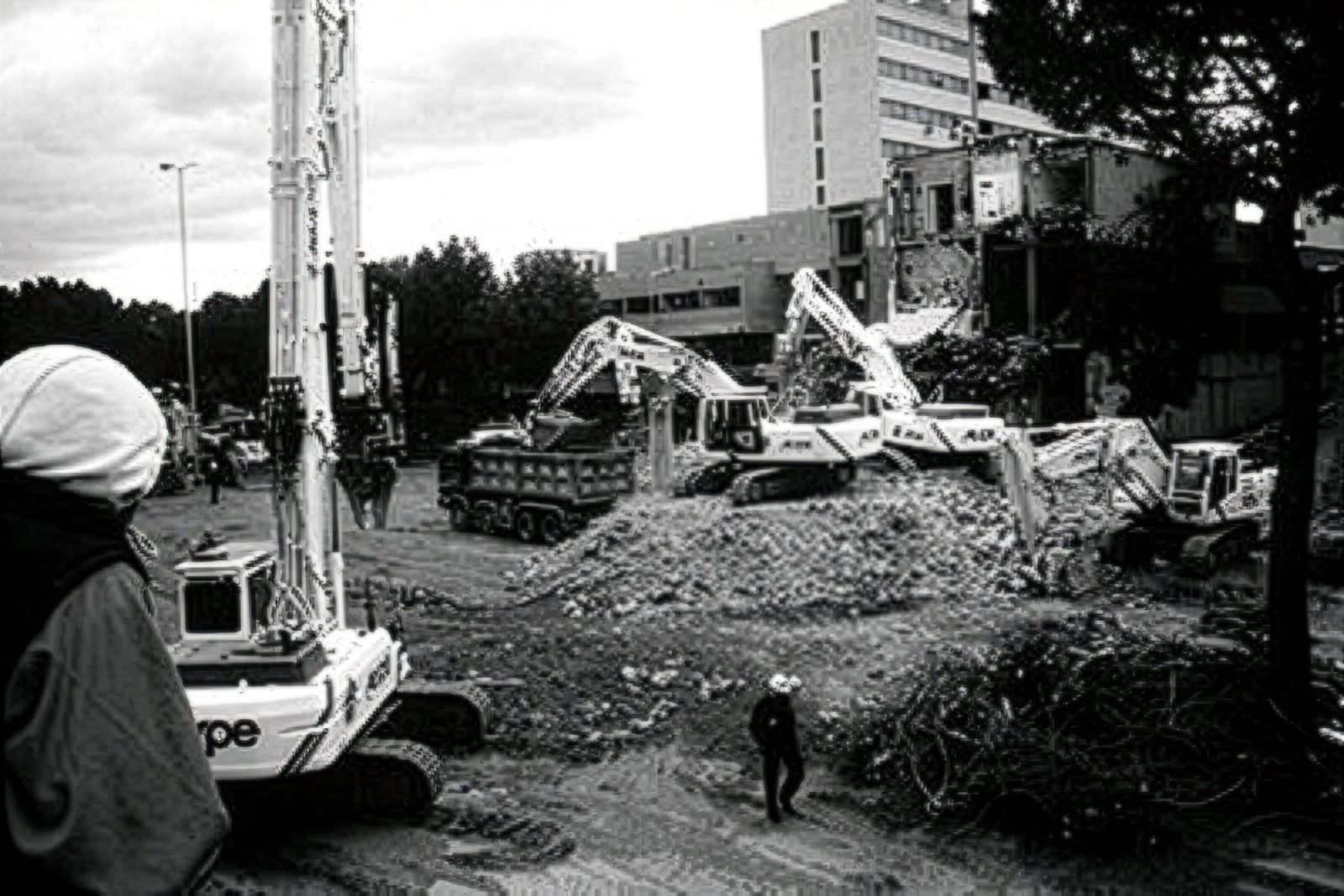 A construction site with multiple excavators moving dirt and rubble, surrounded by buildings and trees, with workers present in the scene.