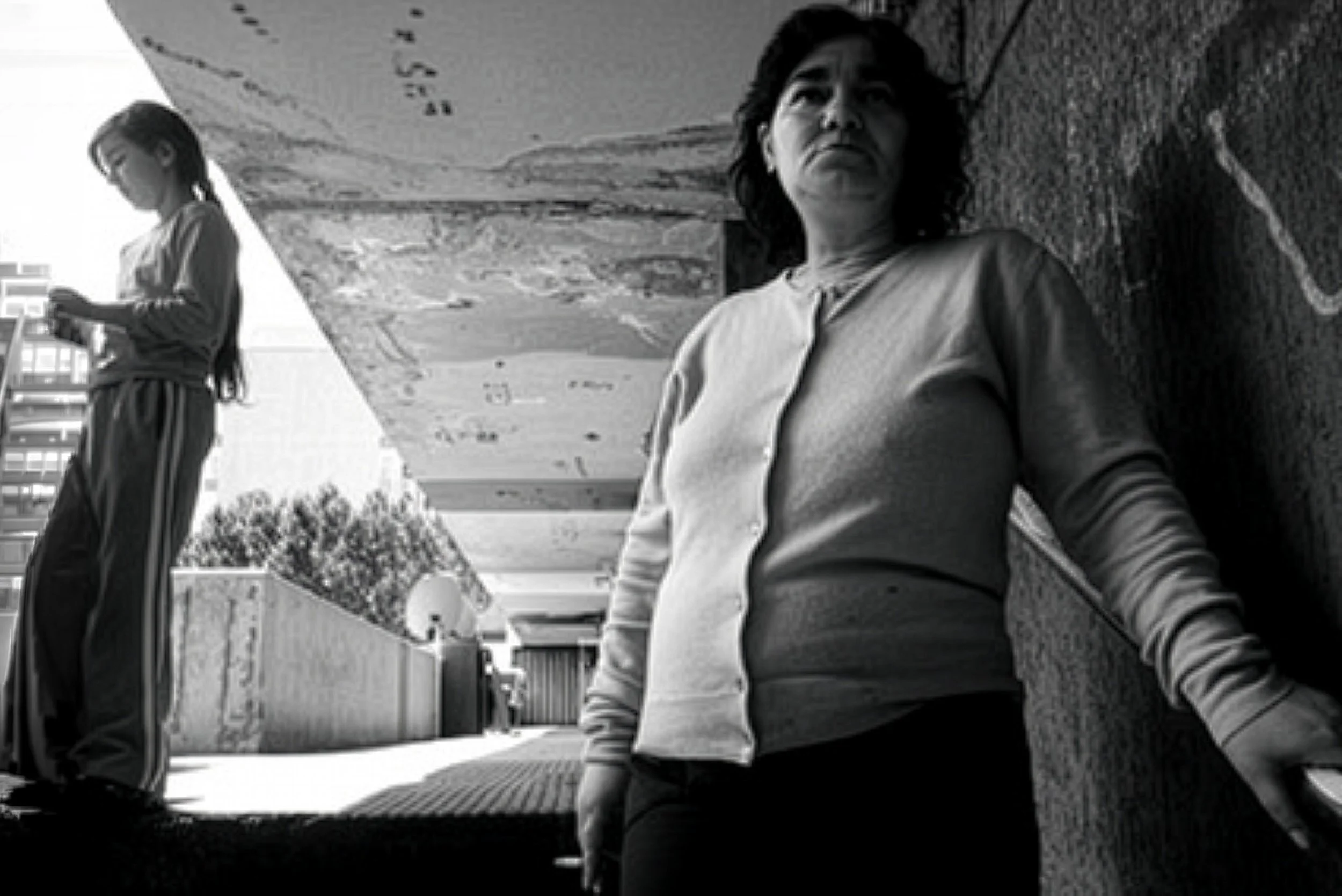 Two women in an outdoor urban setting, with one woman leaning against a wall and the other standing further back looking at her phone.