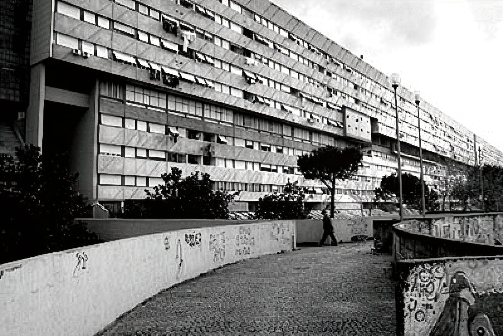 A black and white photo of a large multi-story apartment building with many windows and air conditioning units. In front, there is a curved pathway with street lamps, trees, and a person walking.