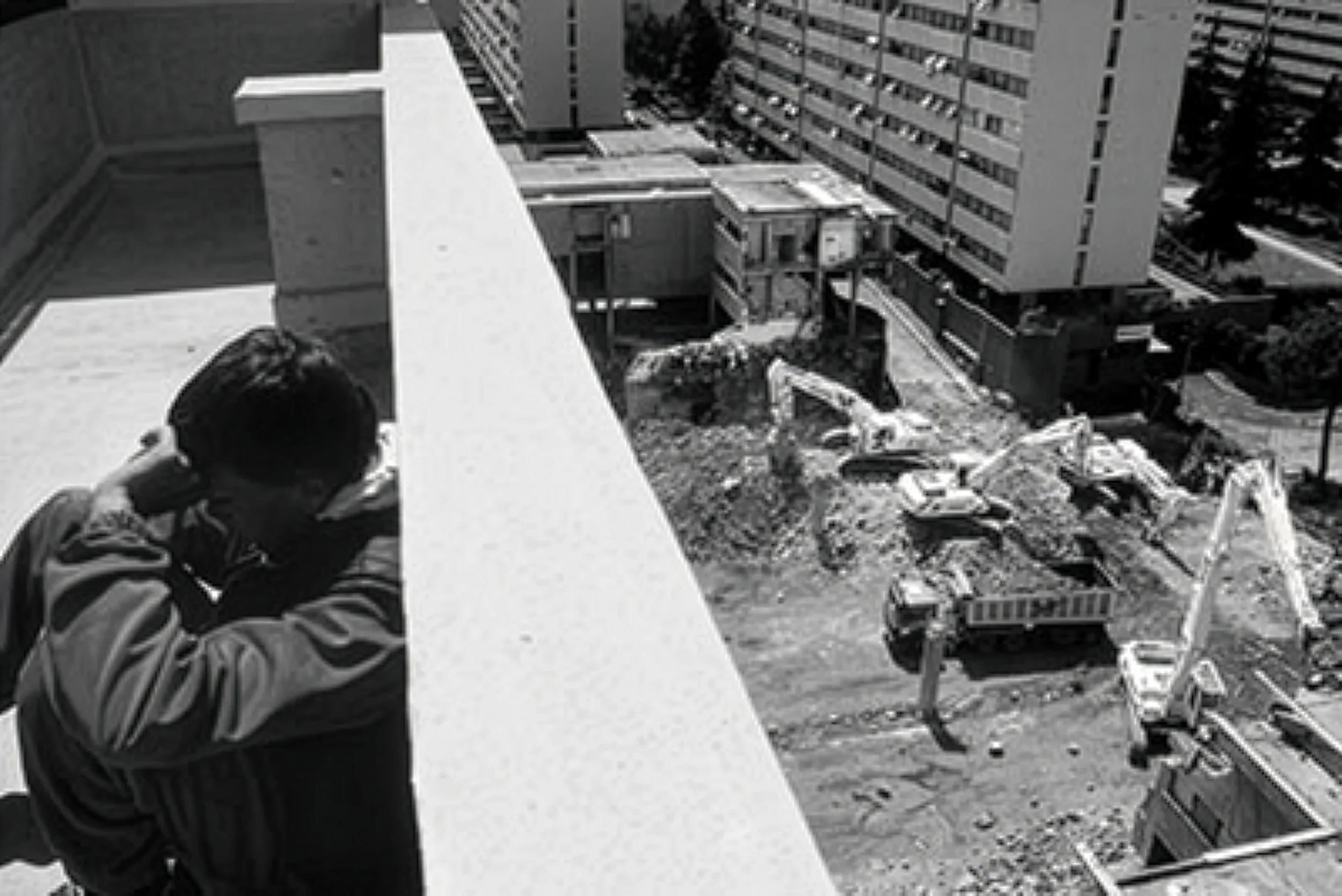 A person standing on a balcony looks down at a construction site with excavators and construction vehicles in a cityscape with tall buildings.