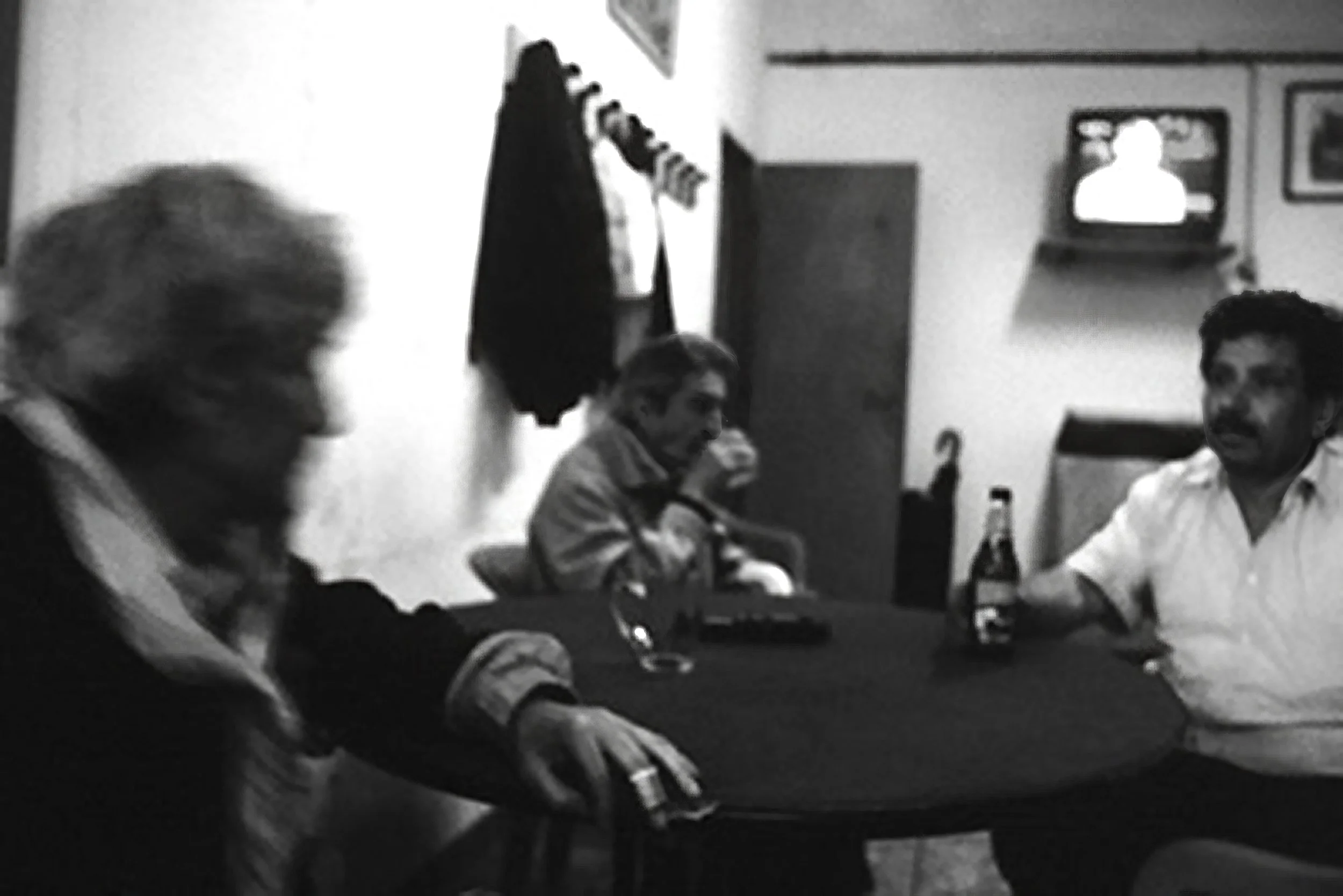 Three people sitting around a table in a room, with a woman in the foreground on the left, a man in the middle, and another man on the right. There are bottles and glasses on the table, and a TV mounted on the wall in the background.