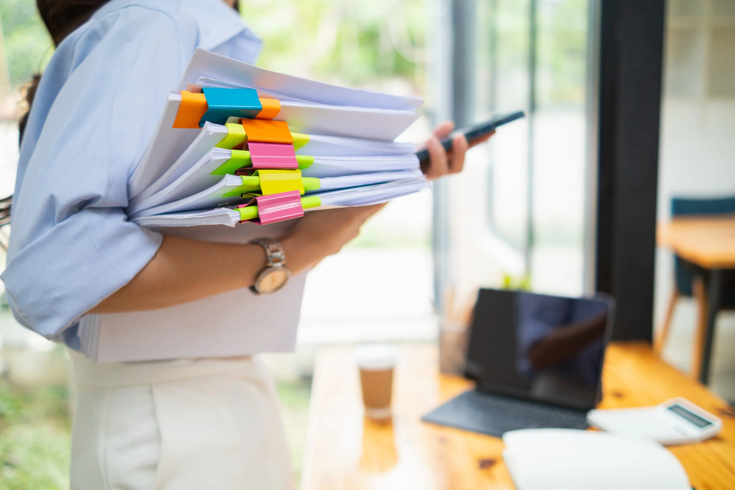 Person holding a stack of papers clipped with colorful binder clips, holding a smartphone, with a laptop and coffee on a wooden table nearby.