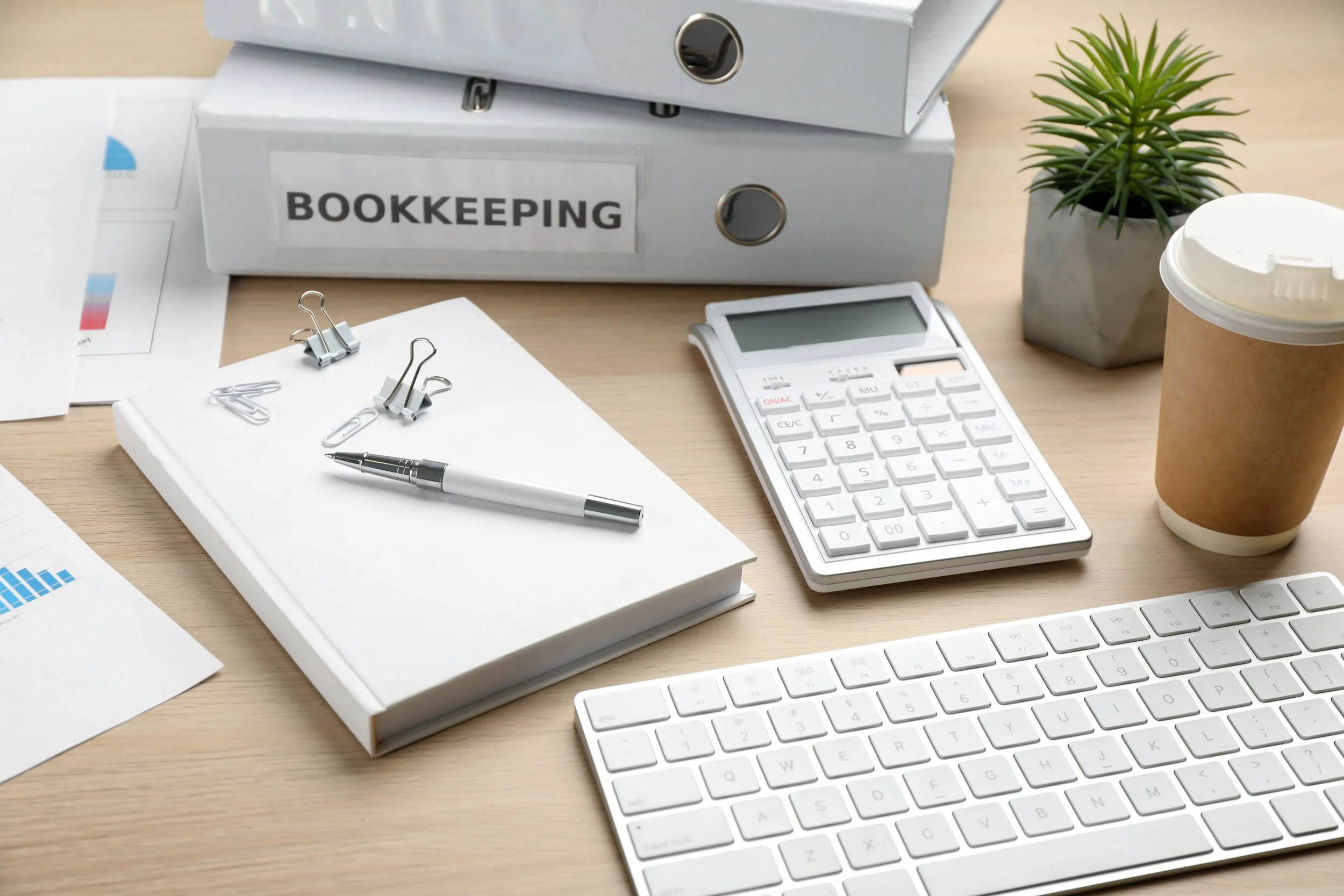 Office desk with bookkeeping materials, calculator, keyboard, pen, paper clips, binders, small plant, coffee cup.