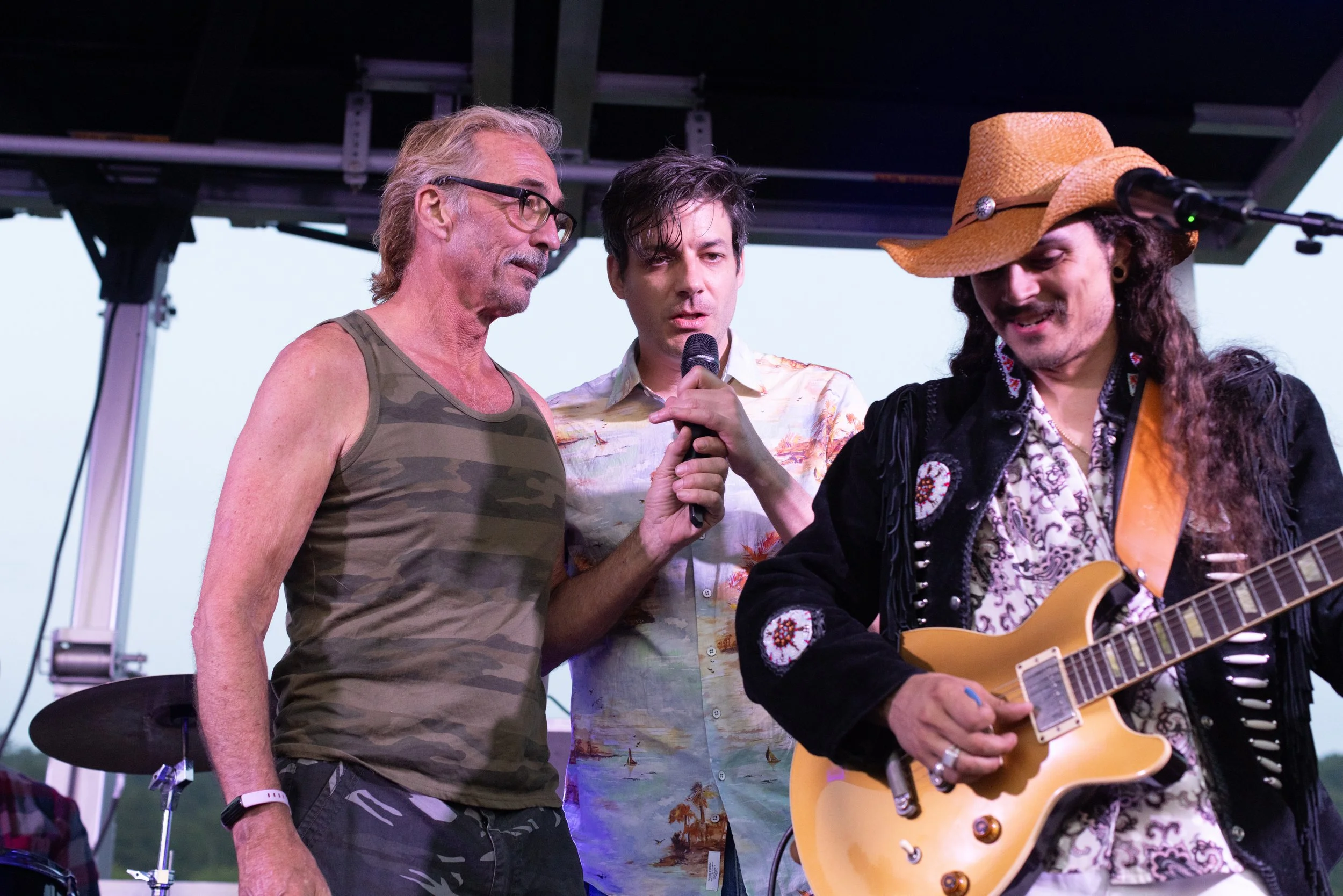 Three men on stage, one playing guitar, another holding a microphone, and the third speaking. The man on the left has gray hair, glasses, and a sleeveless camouflage shirt. The man in the middle has dark hair and is wearing a button-up shirt with a b