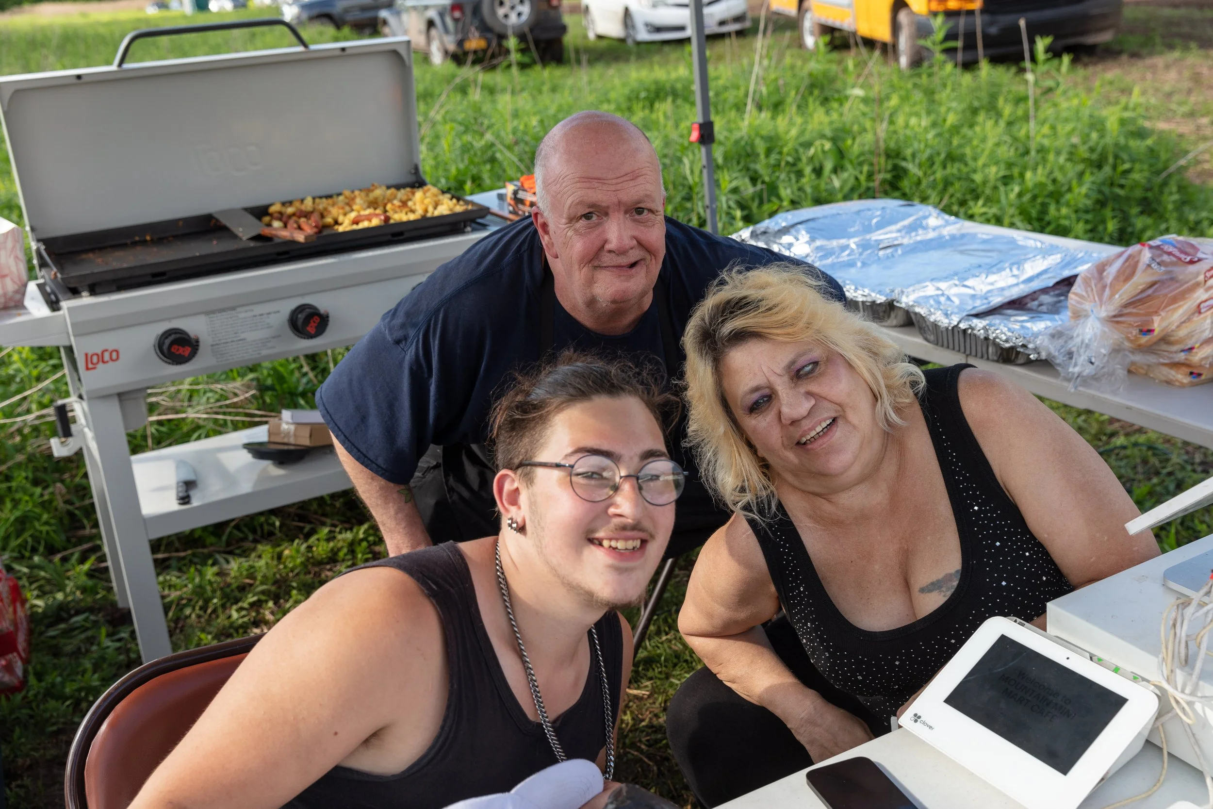 A group of four people, two women and two men, smiling and posing outdoors in front of a portable grill with food, during daytime. There are green fields and parked cars in the background.