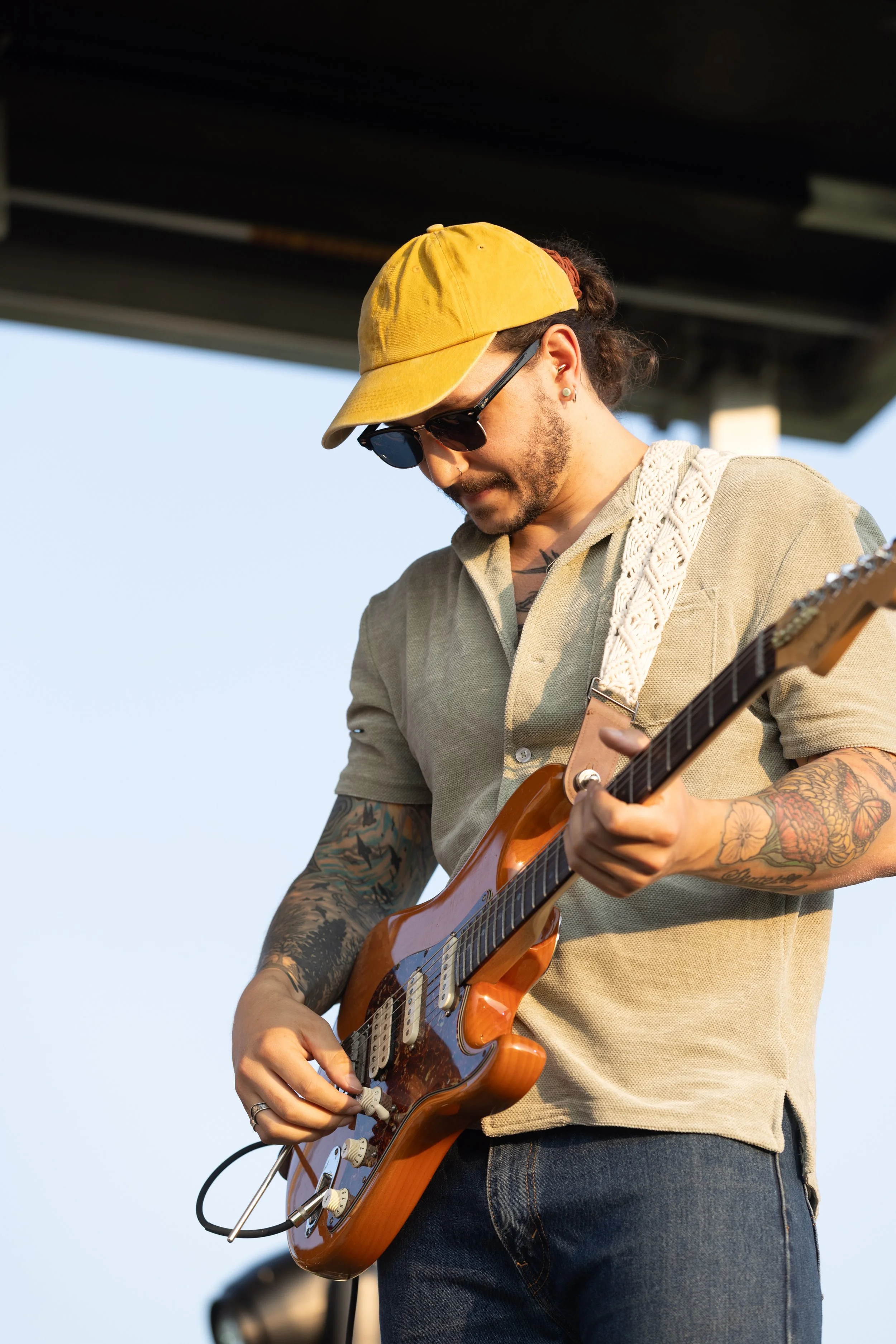 A man with tattoos on his arms, wearing a yellow cap, sunglasses, and a beige shirt, playing an electric guitar outdoors under a bridge or overpass during daylight.