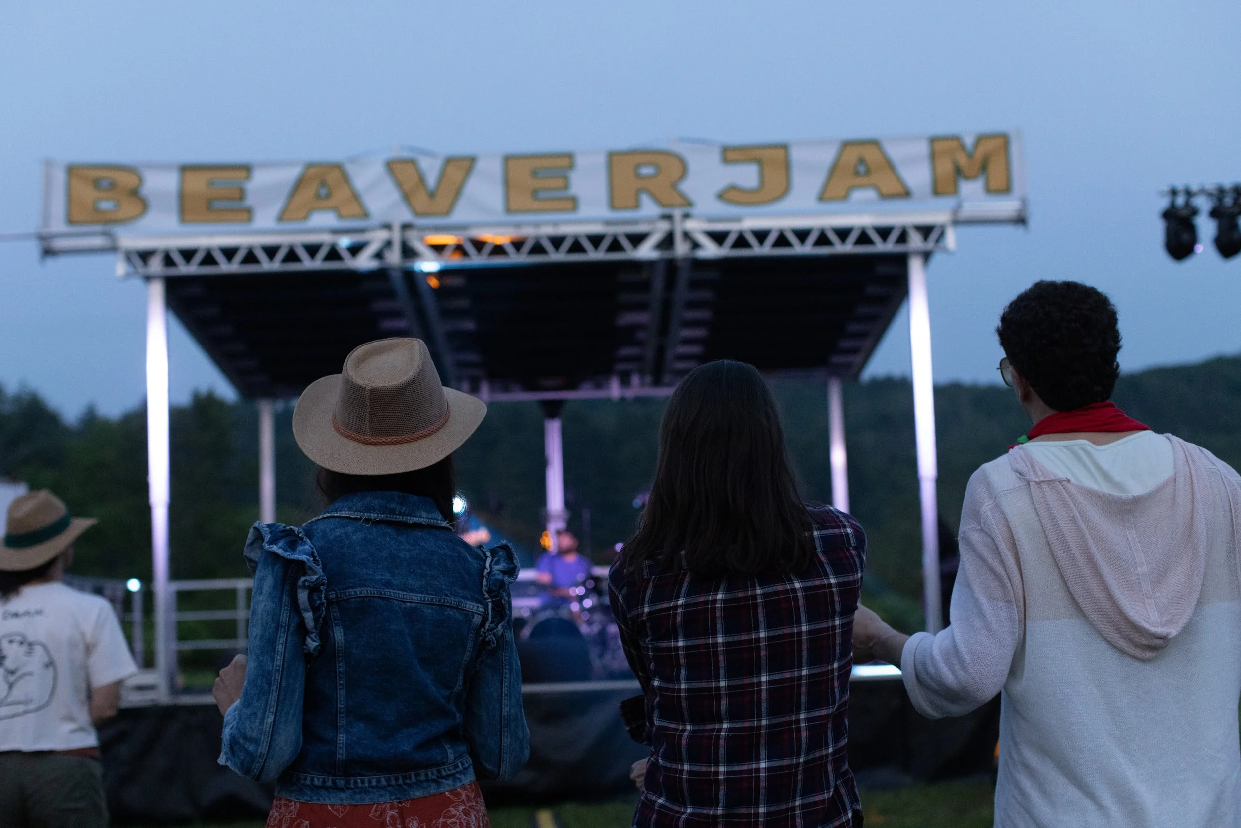 Group of people watching an outdoor concert or event during dusk, with a large sign reading 'BEAVER JAM' above the stage.