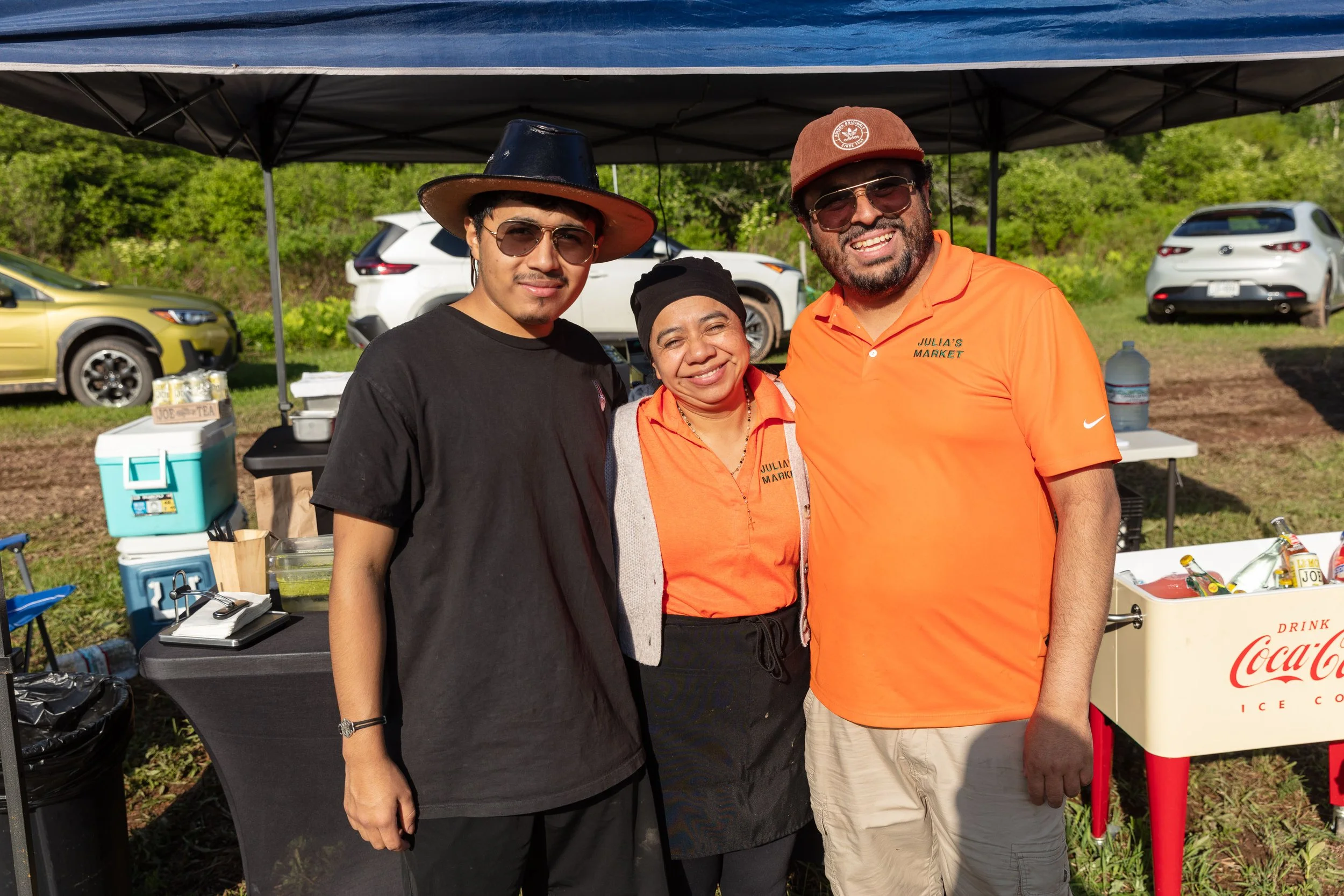 Three people standing together under a canopy at an outdoor event, smiling at the camera. Two men wearing sunglasses and hats, and a woman in the middle wearing a black headscarf and an orange shirt with a name tag. Cars and greenery are in the backg