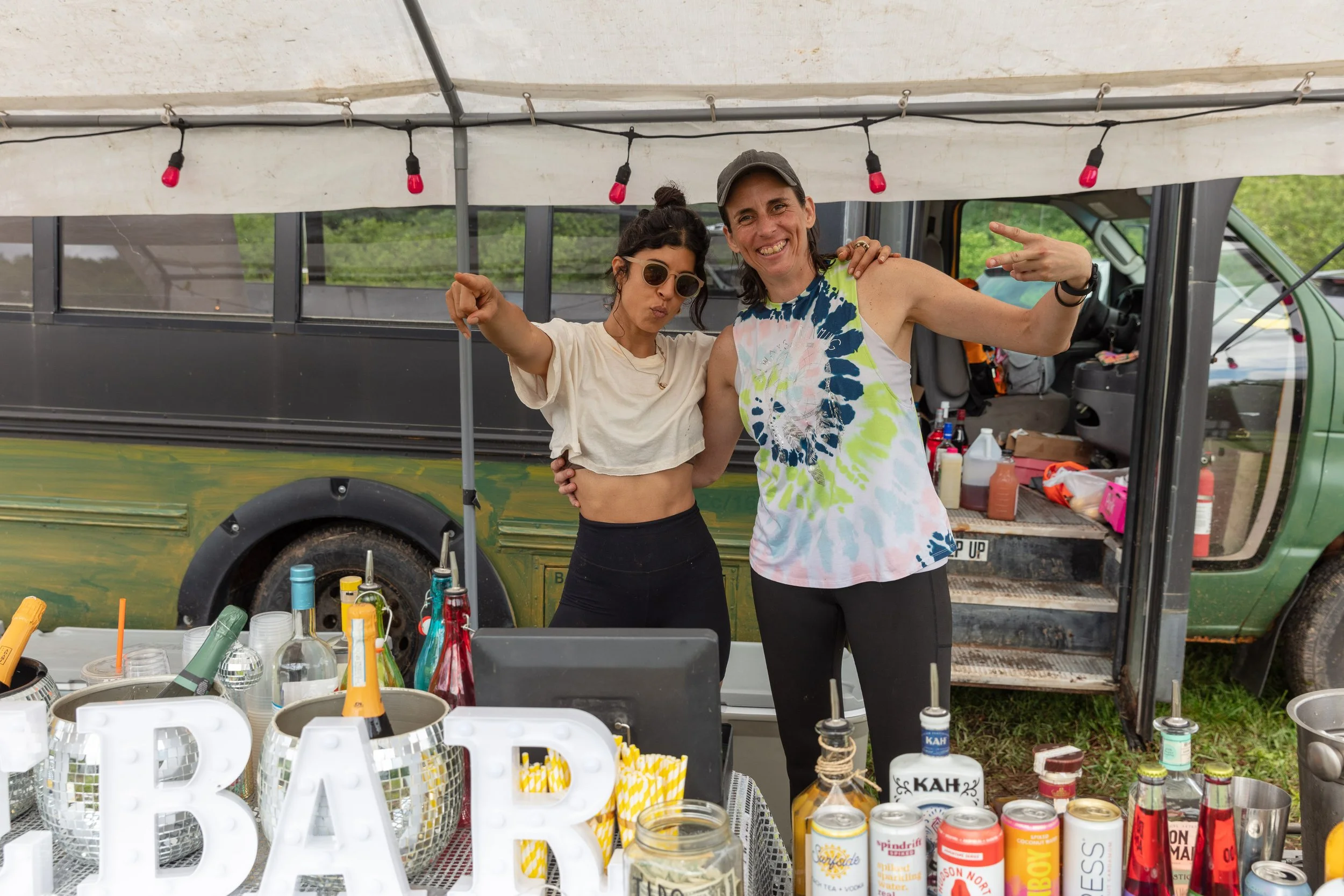 Two women are posing happily at an outdoor event stand under a canopy with string lights. They are surrounded by various bottles, cans, and decorations, with a green bus in the background.