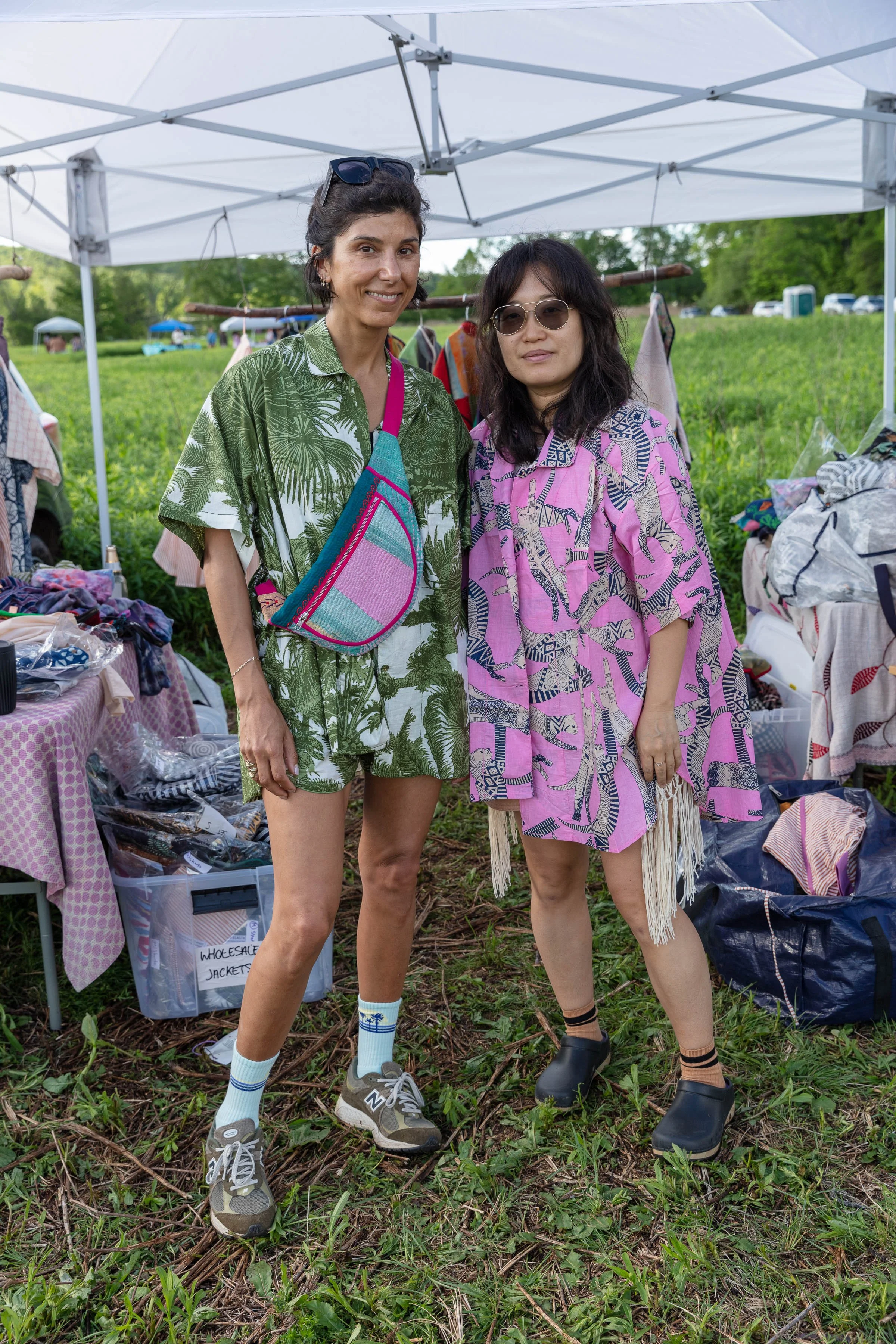 Two women standing under a white canopy at an outdoor market, surrounded by clothing and accessories, with green trees and a grassy field in the background.