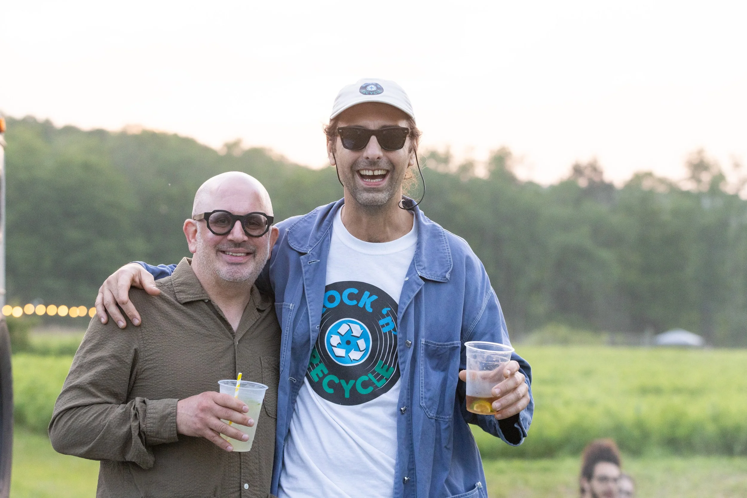 Two men smiling and holding drinks at an outdoor event during sunset. One man has a beard, glasses, and is wearing a brown jacket. The other man has a beard, sunglasses, a white cap, and a blue jacket. They are standing close, with the background of 