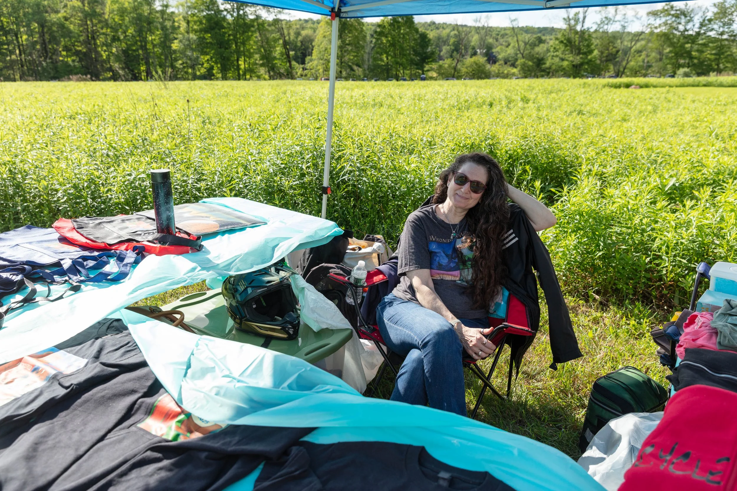 A woman with long curly hair wearing sunglasses, a dark t-shirt, and blue jeans sitting in a foldable chair outdoors next to a table covered with various items, under a blue canopy tent. The background shows a lush green field of tall grass and trees