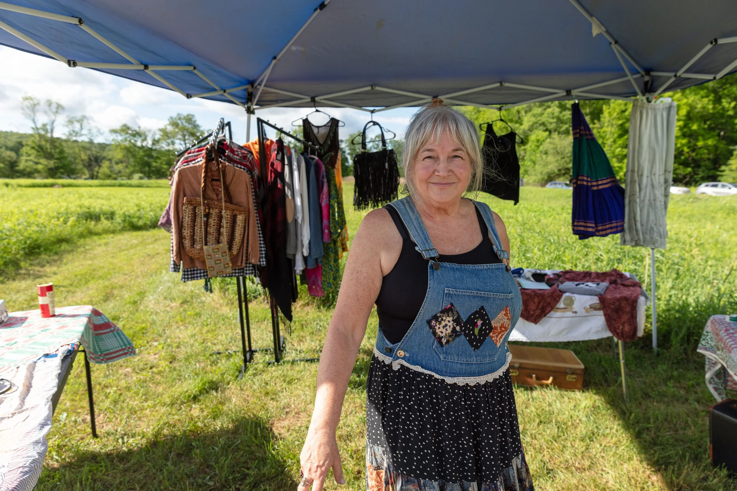 A woman with gray hair smiling at an outdoor market stall under a blue canopy, with green fields and trees in the background. The stall displays clothing on hangers and tables with fabric and other items.