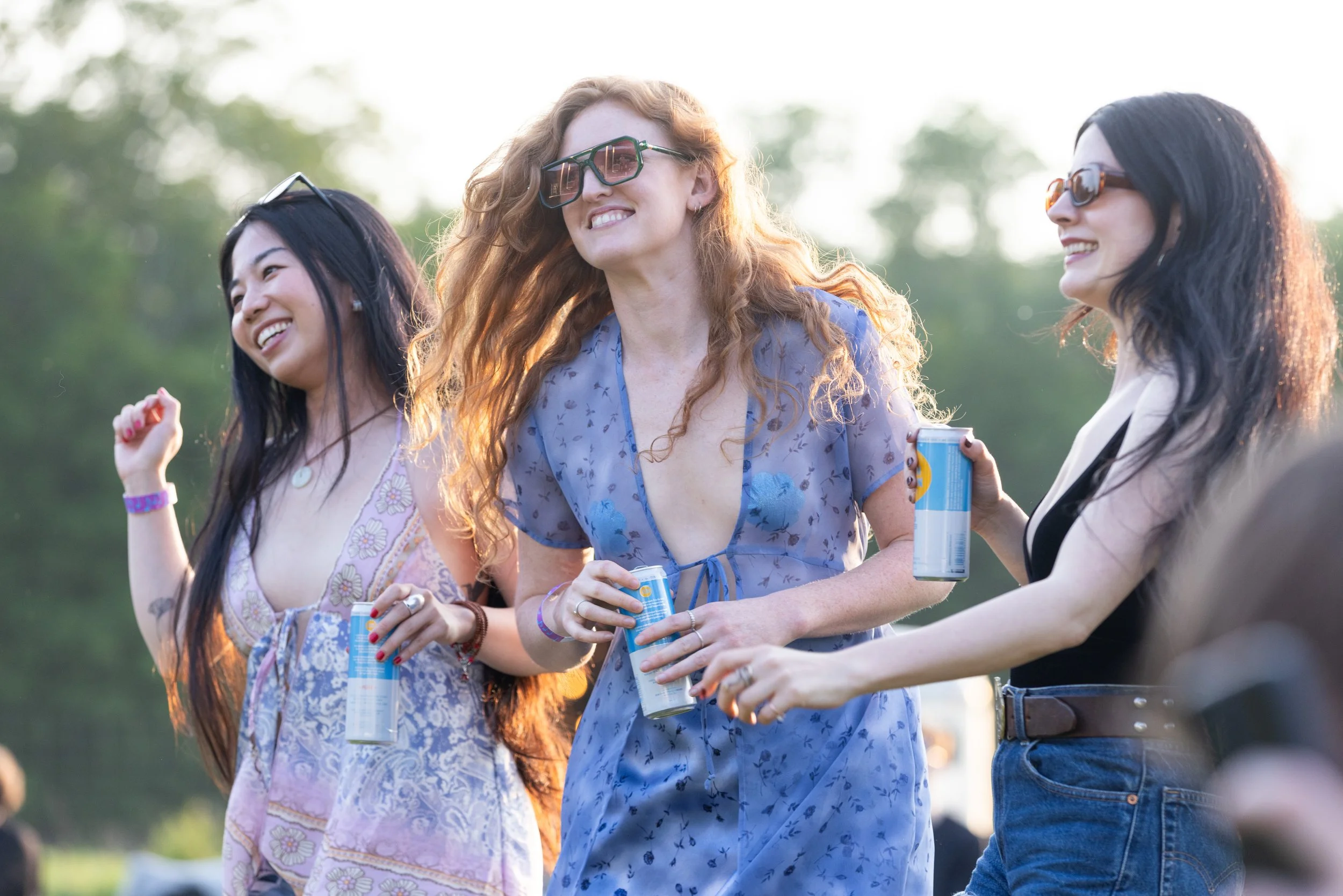 Three women smiling and enjoying drinks outdoors, wearing sunglasses, with trees and sunlight in the background.