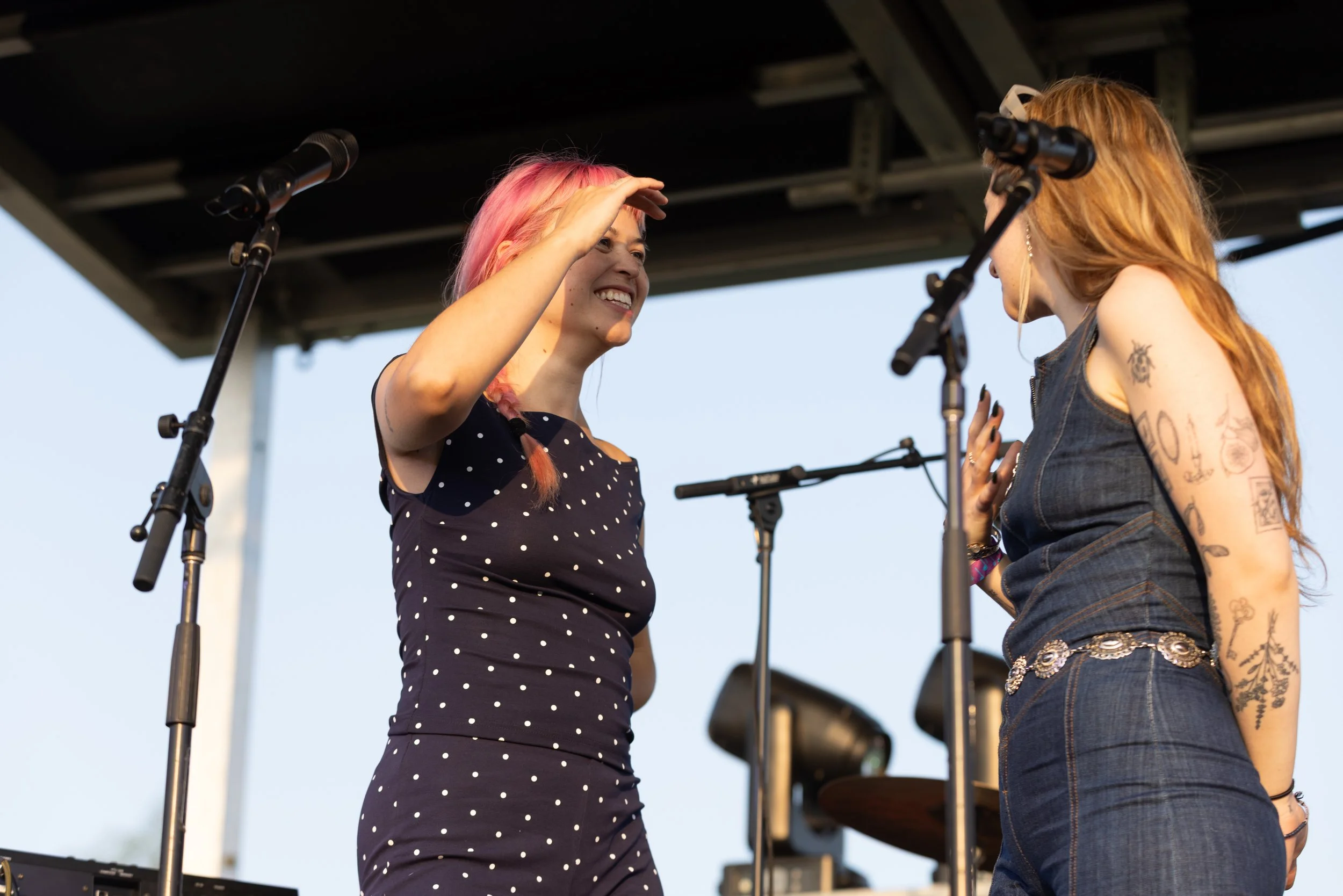 Two women on a stage near microphones, engaging in conversation, with one woman smiling and the other speaking or listening, outdoors during sunset.