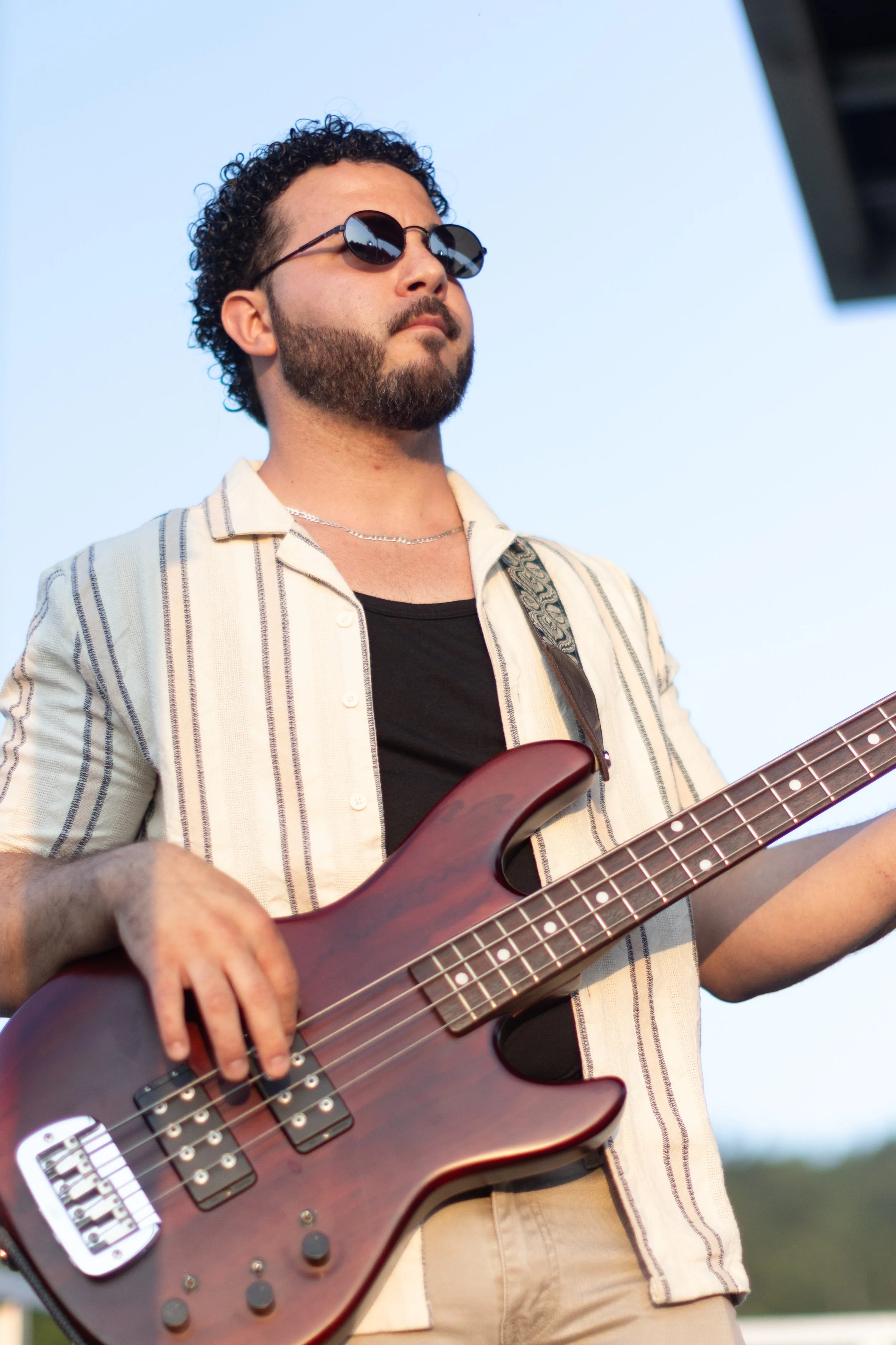 A man with dark curly hair, sunglasses, and a beard is playing a brown electric bass guitar outdoors against a blue sky.