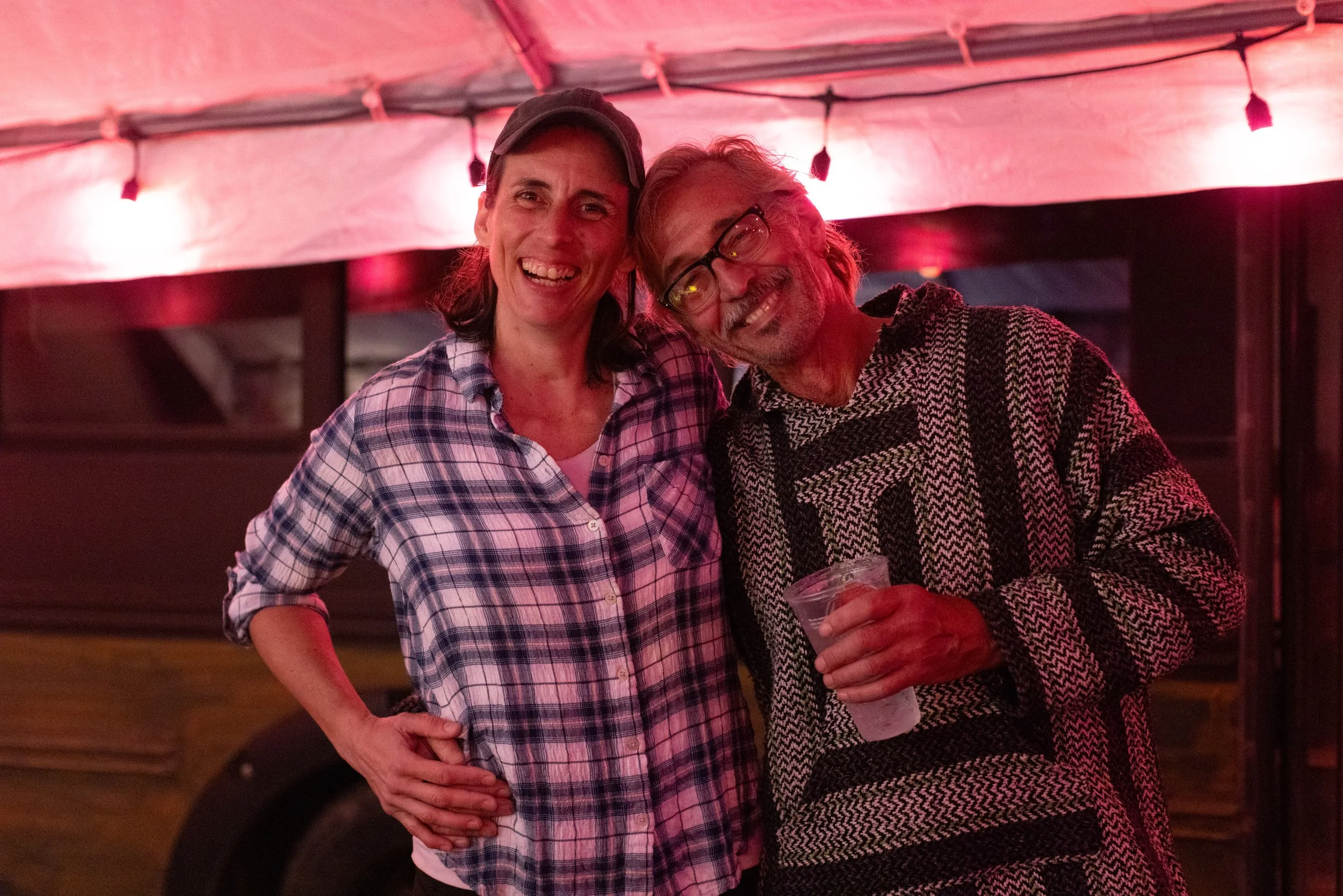 A woman and a man are smiling and standing close together in a dimly lit room with red lighting. The woman is wearing a plaid shirt and a baseball cap, and the man is wearing glasses and a patterned sweater while holding a drink.