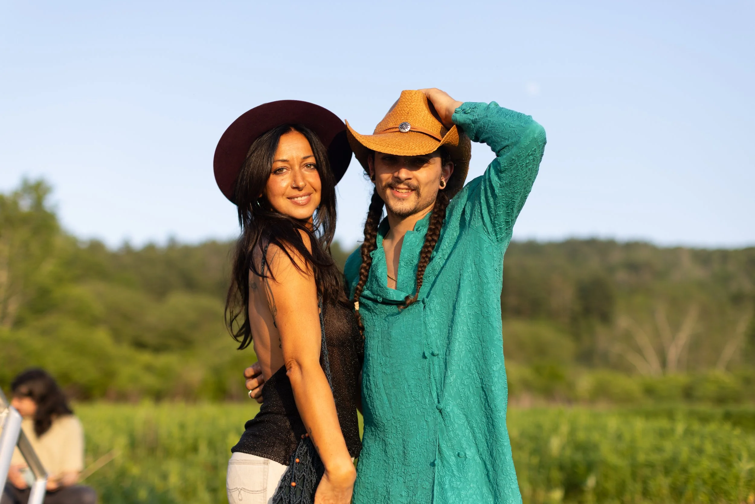 Two people are standing outdoors during daytime, both wearing wide-brimmed hats. One person has long dark hair and is dressed in a black top and white pants. The other person has long braids and is wearing a teal shirt. They are smiling and looking a