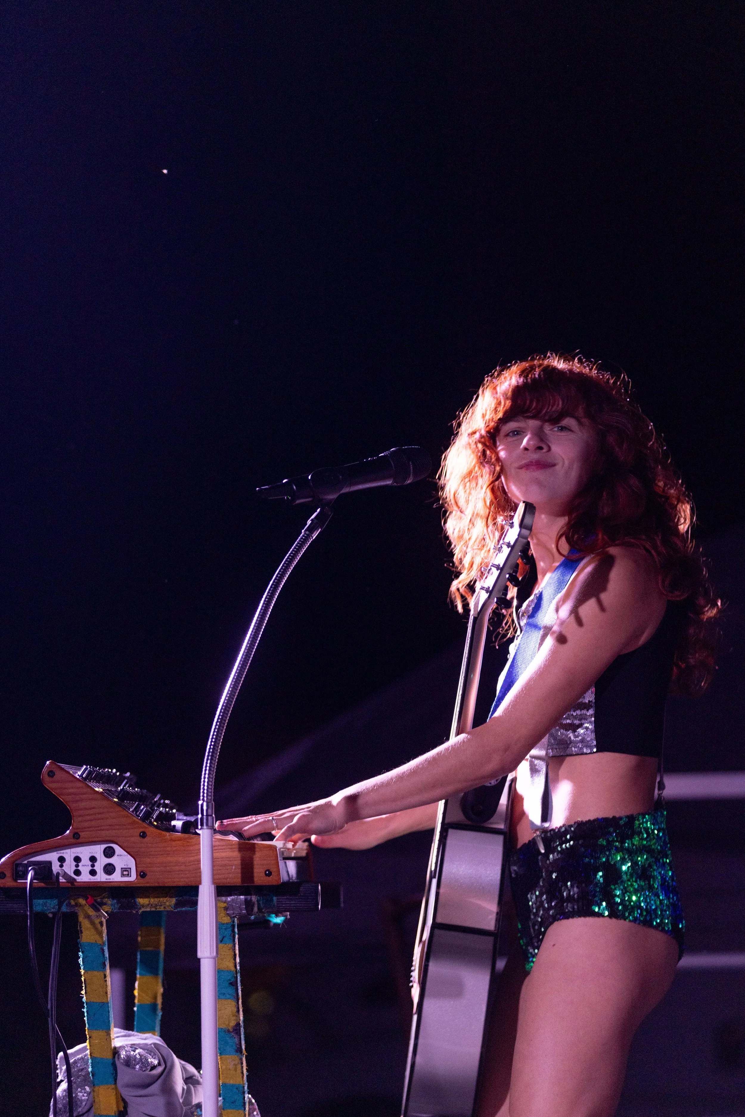 A young woman with curly red hair playing a keyboard on stage, wearing a shiny black top, green sparkly shorts, and standing in front of a microphone, with a dark background.