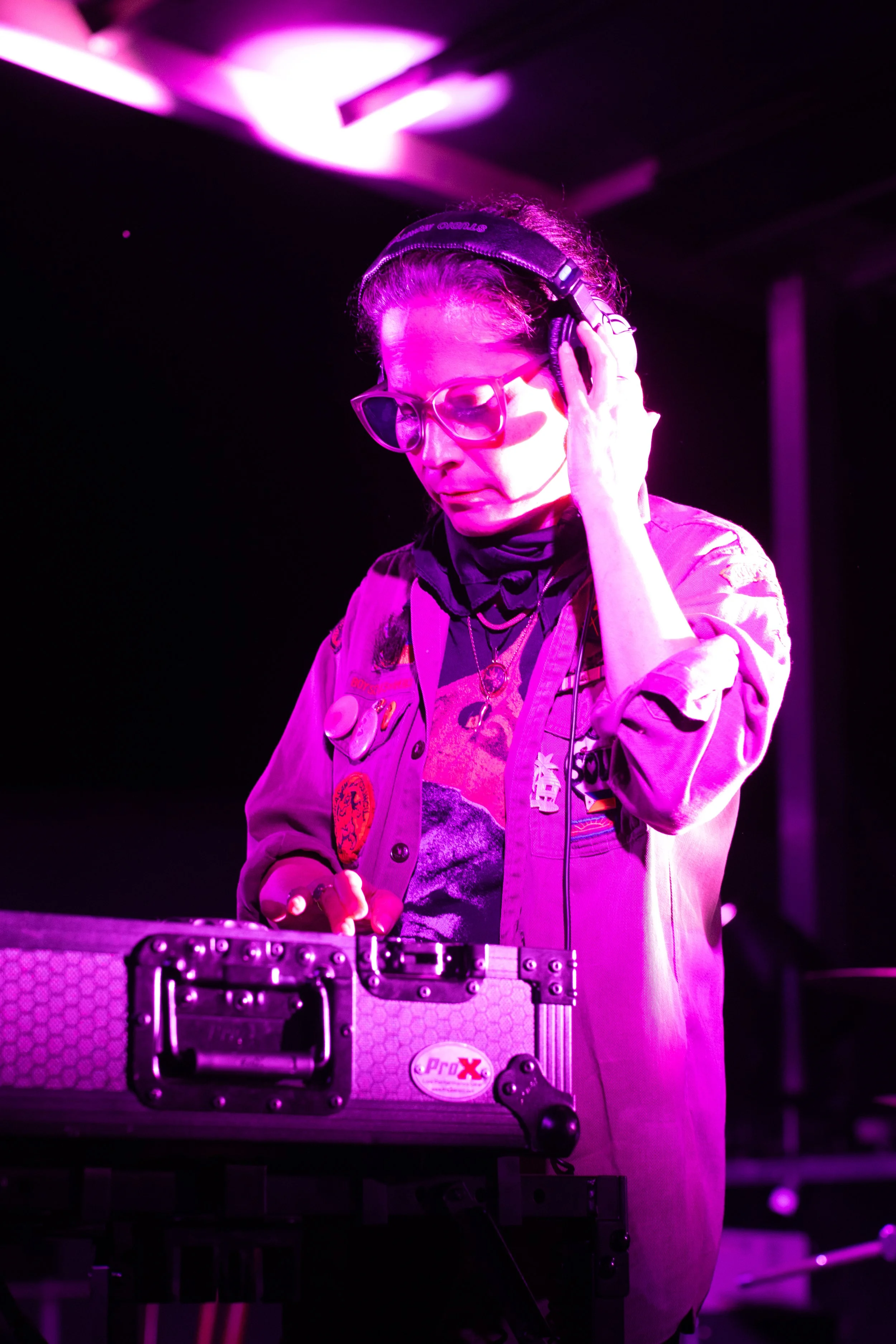 A woman with glasses and a bandana on her head is DJing, with pink and purple stage lighting illuminating her. She is adjusting headphones while manipulating DJ equipment.
