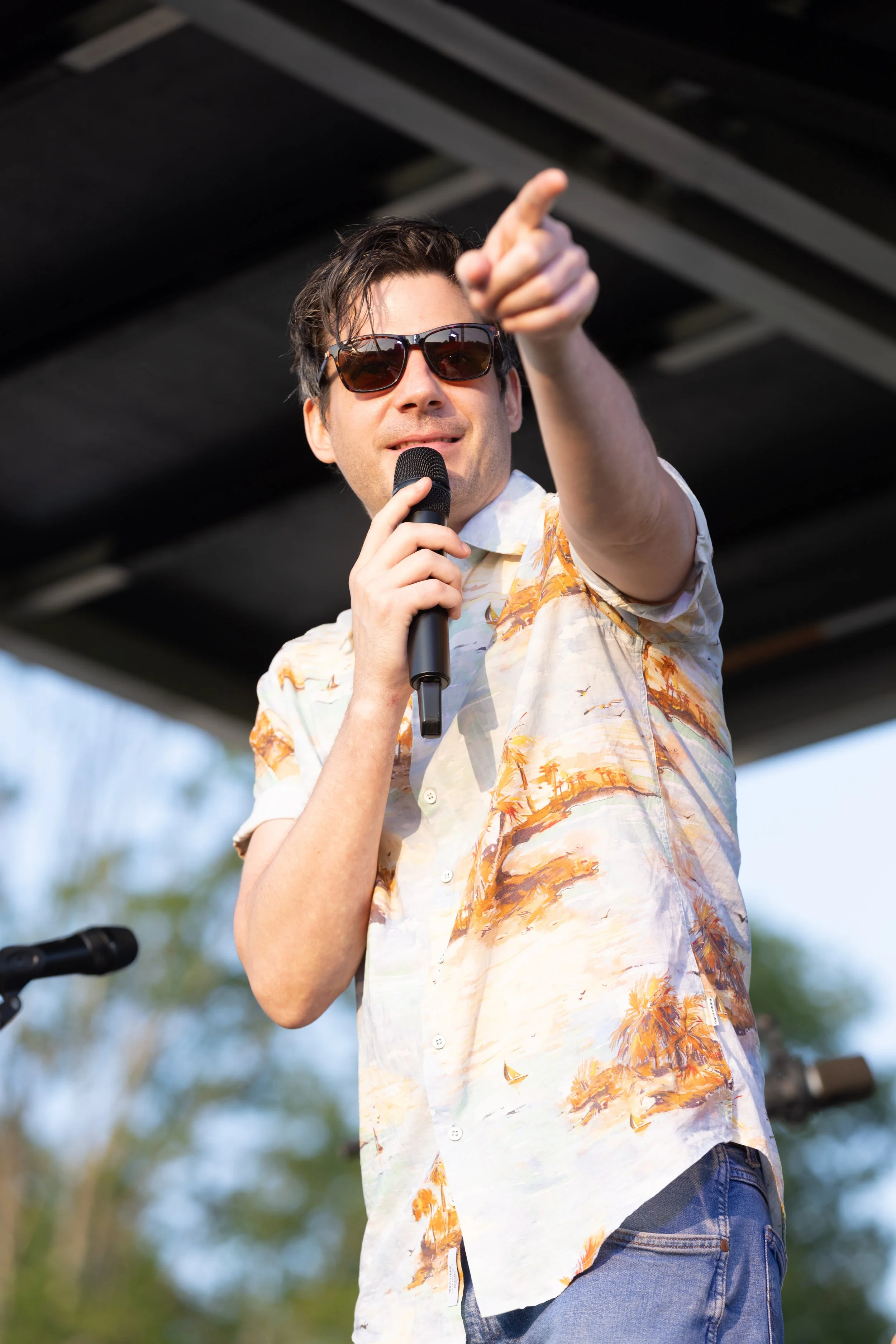A man wearing sunglasses and a floral shirt is speaking into a microphone and pointing outward, outdoors under a cloudy sky.