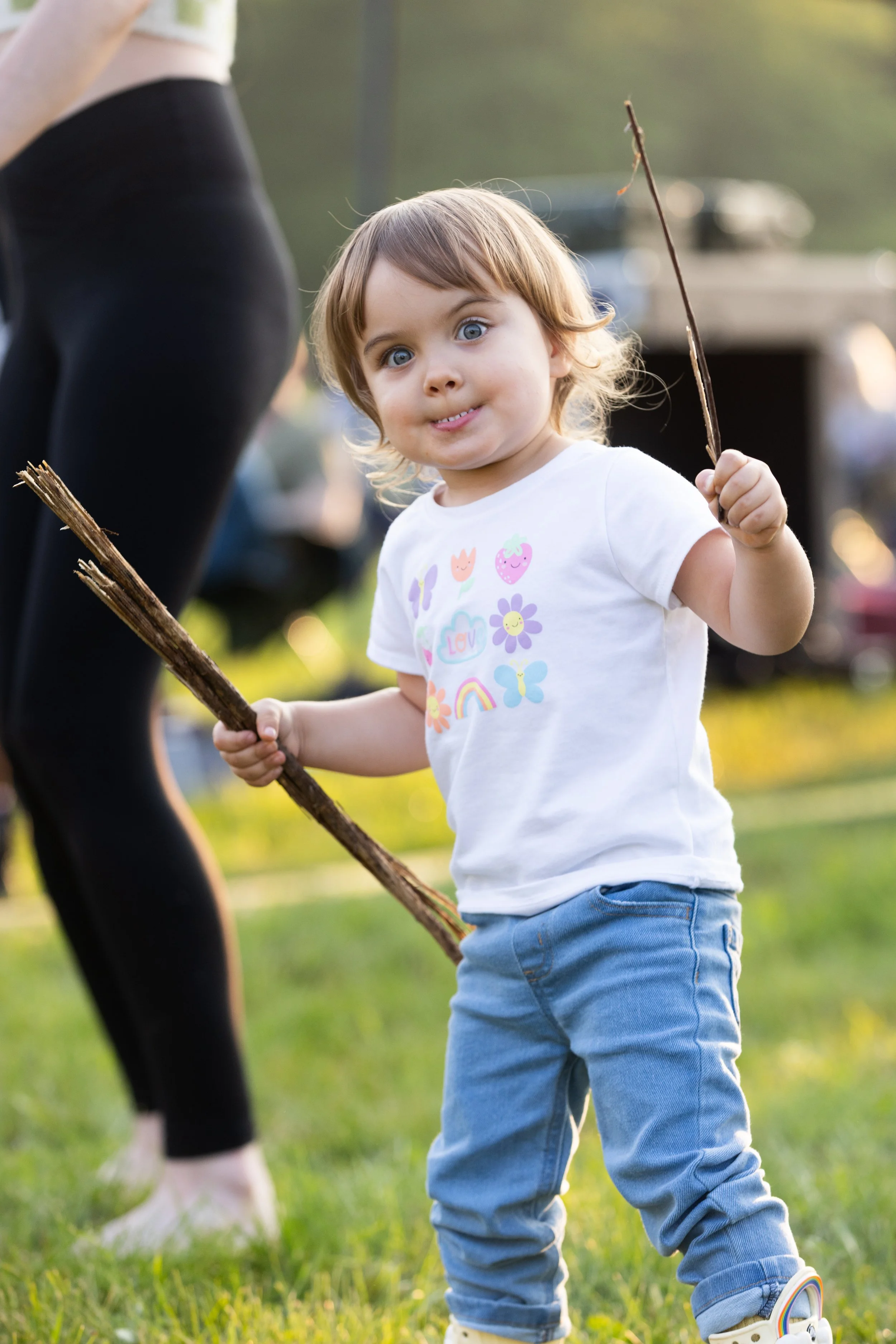 Young child with brown hair and blue eyes holding a stick in a park, wearing a white t-shirt with colorful flowers and blue jeans, with another person seen from the waist down nearby.