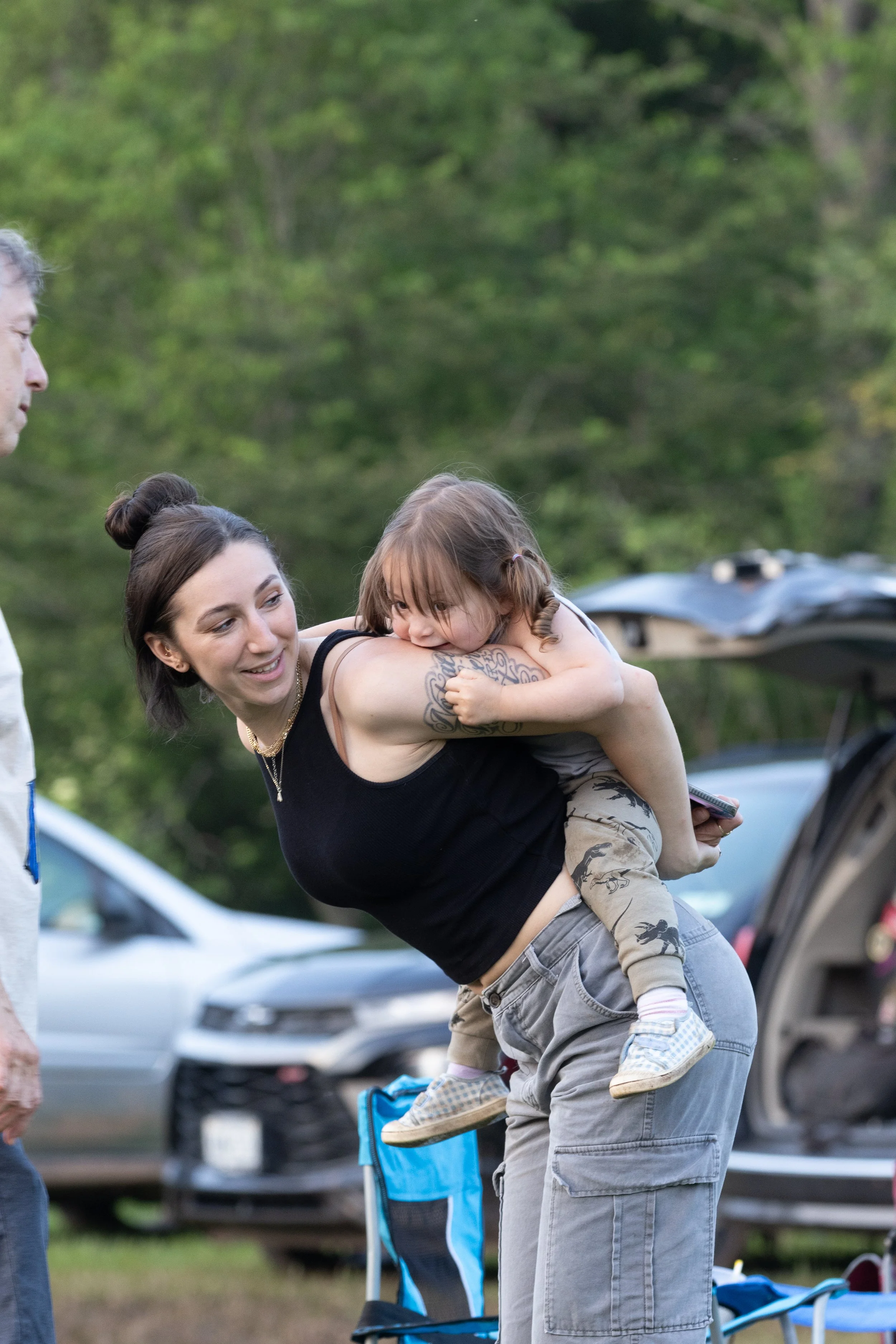 A woman holding a young girl on her back outdoors during daytime, with people, cars, and trees in the background.