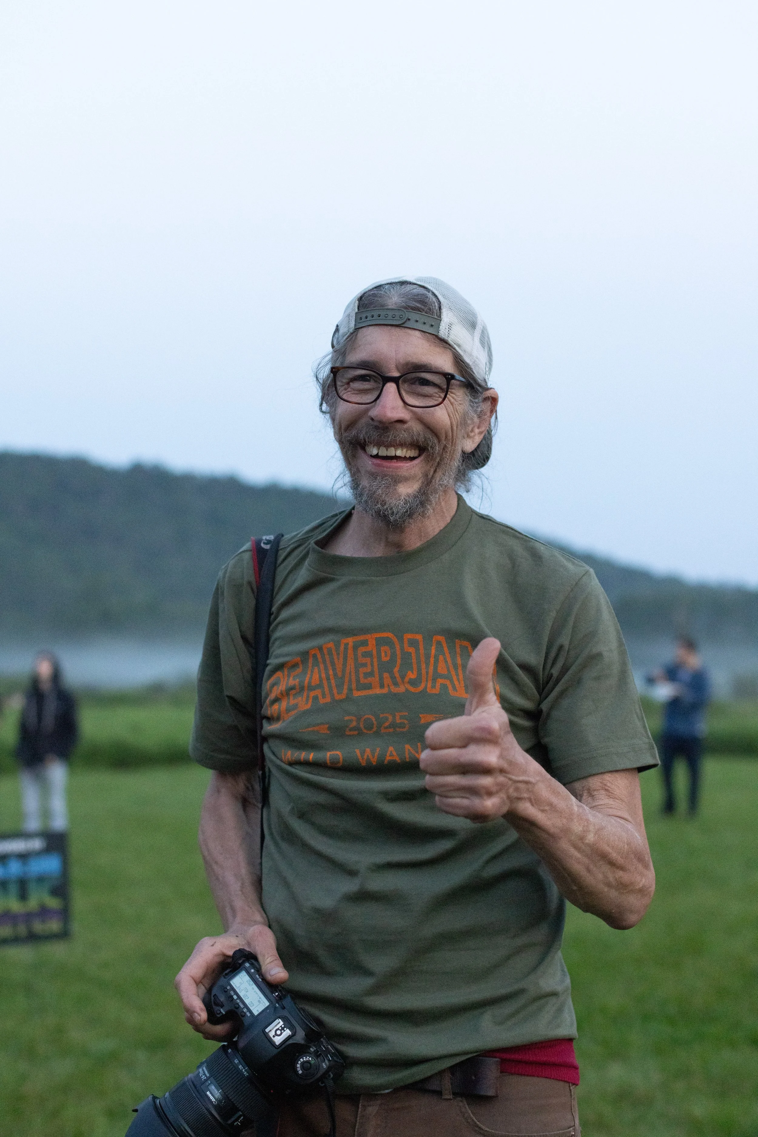 A joyful man with glasses and a backward cap giving a thumbs-up while holding a camera, outdoors with a blurred background of a group of people and a hilly landscape in the evening.
