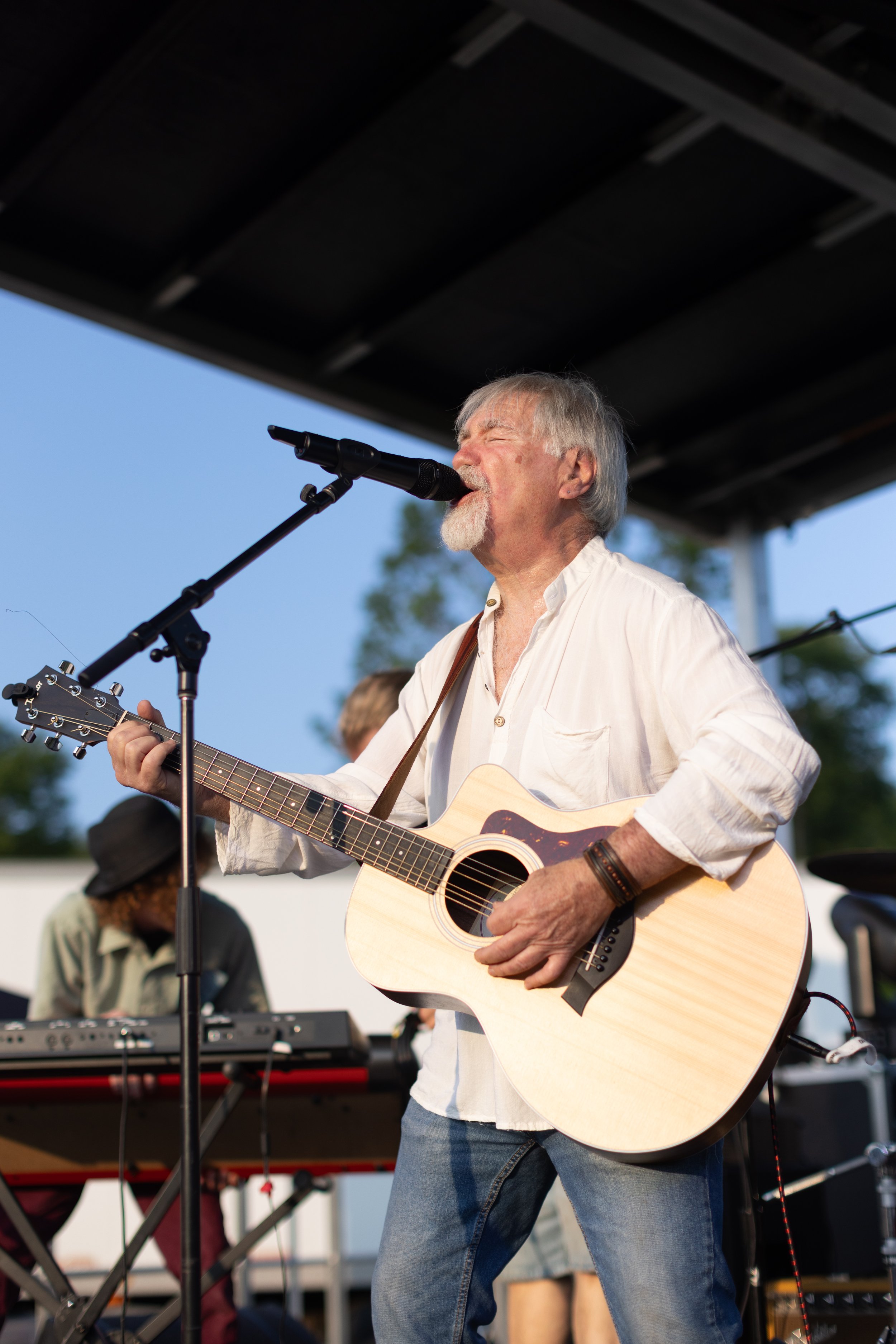 A man with gray hair and a beard playing an acoustic guitar, singing into a microphone outdoors during daylight, with a keyboard player and a person in a hat in the background.