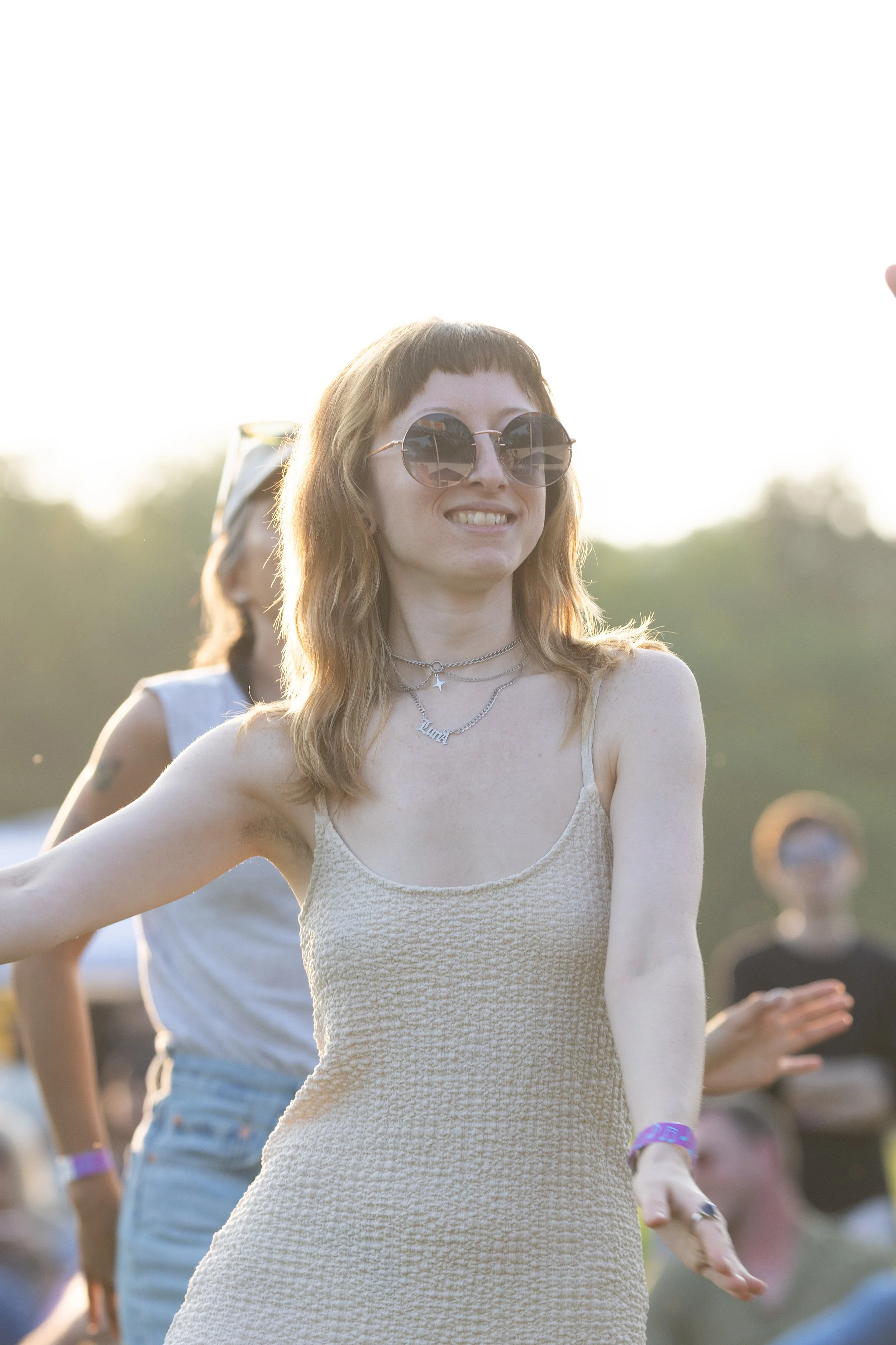 A young woman with light brown hair, wearing sunglasses and a beige sleeveless dress, smiling outdoors during daylight with people in the background.