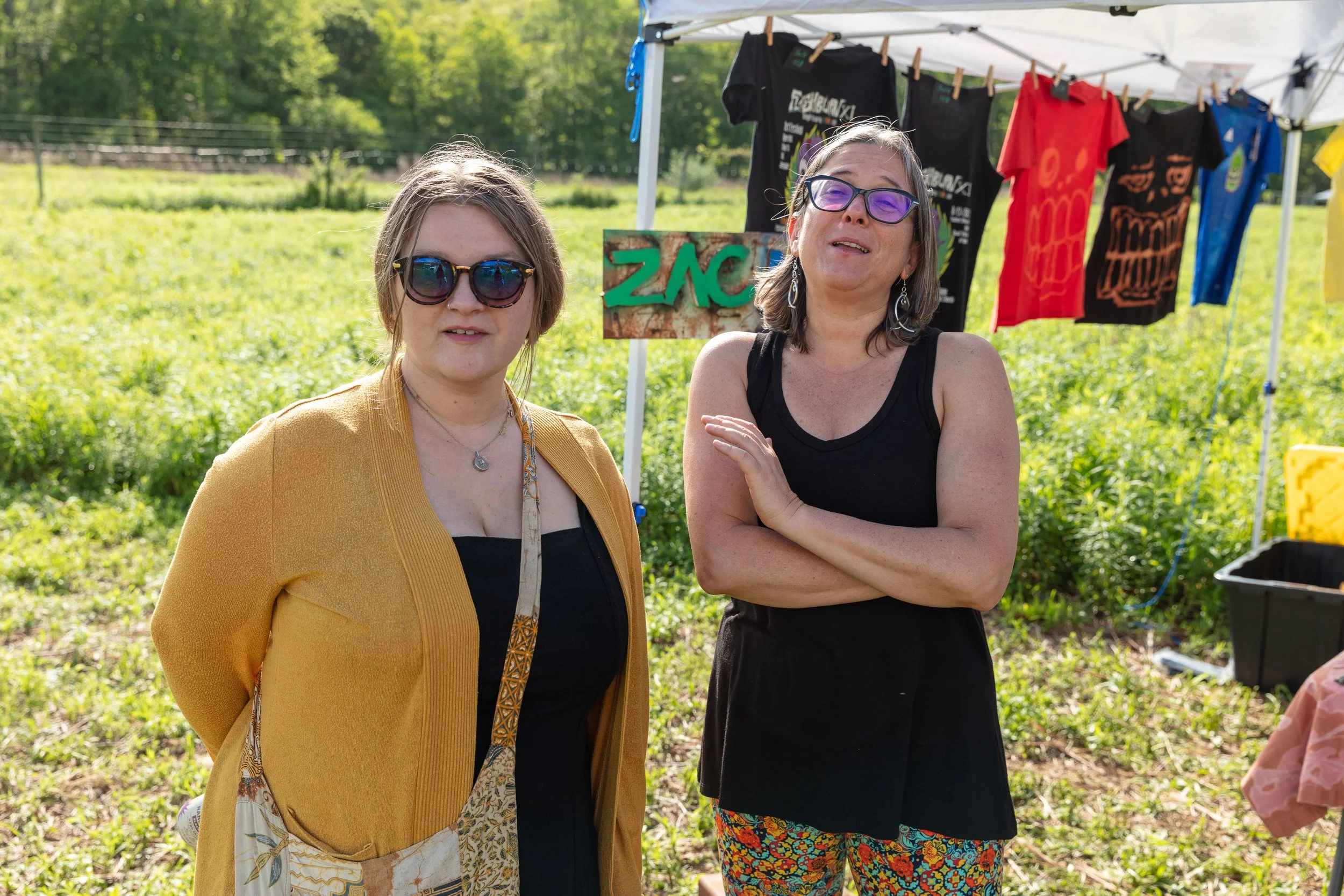 Two women standing outdoors near a white tent with colorful T-shirts hanging on a line behind them. One woman has short hair, sunglasses, and a yellow blazer, while the other has shoulder-length hair, glasses, and a black top.