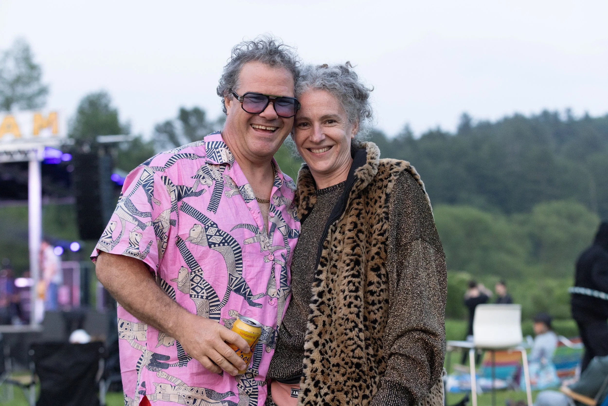 Two smiling people, a man wearing sunglasses and a pink patterned shirt, and a woman with gray curly hair wearing a leopard print jacket, are standing close together outdoors during daytime.