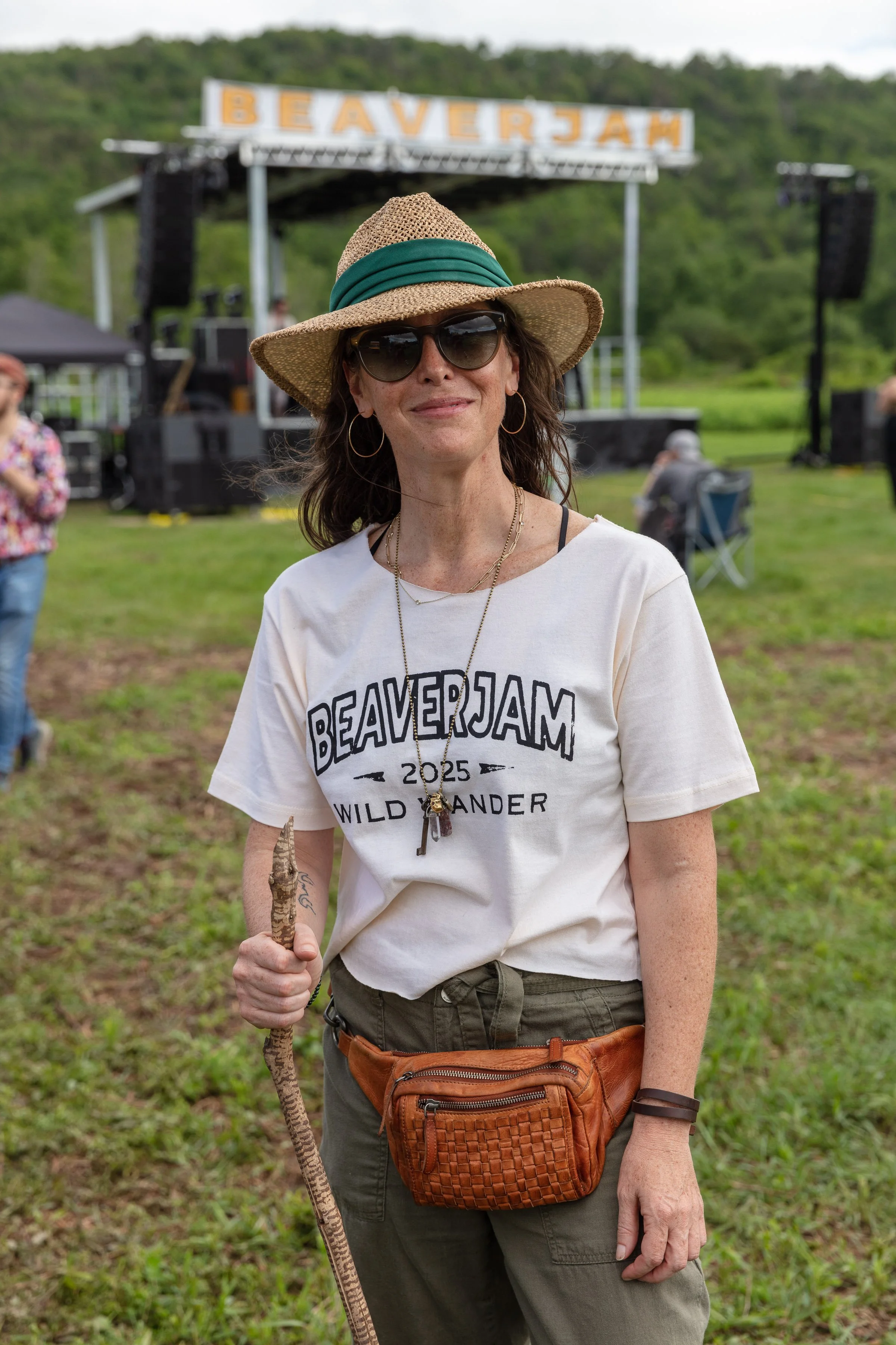 A woman wearing sunglasses, a wide-brimmed hat, and a beige T-shirt with the word 'BEAVERJAM' standing outdoors at an event, holding a walking stick with a carved handle. In the background, there is a stage with a sign that reads 'BEAVERJAM' and peop