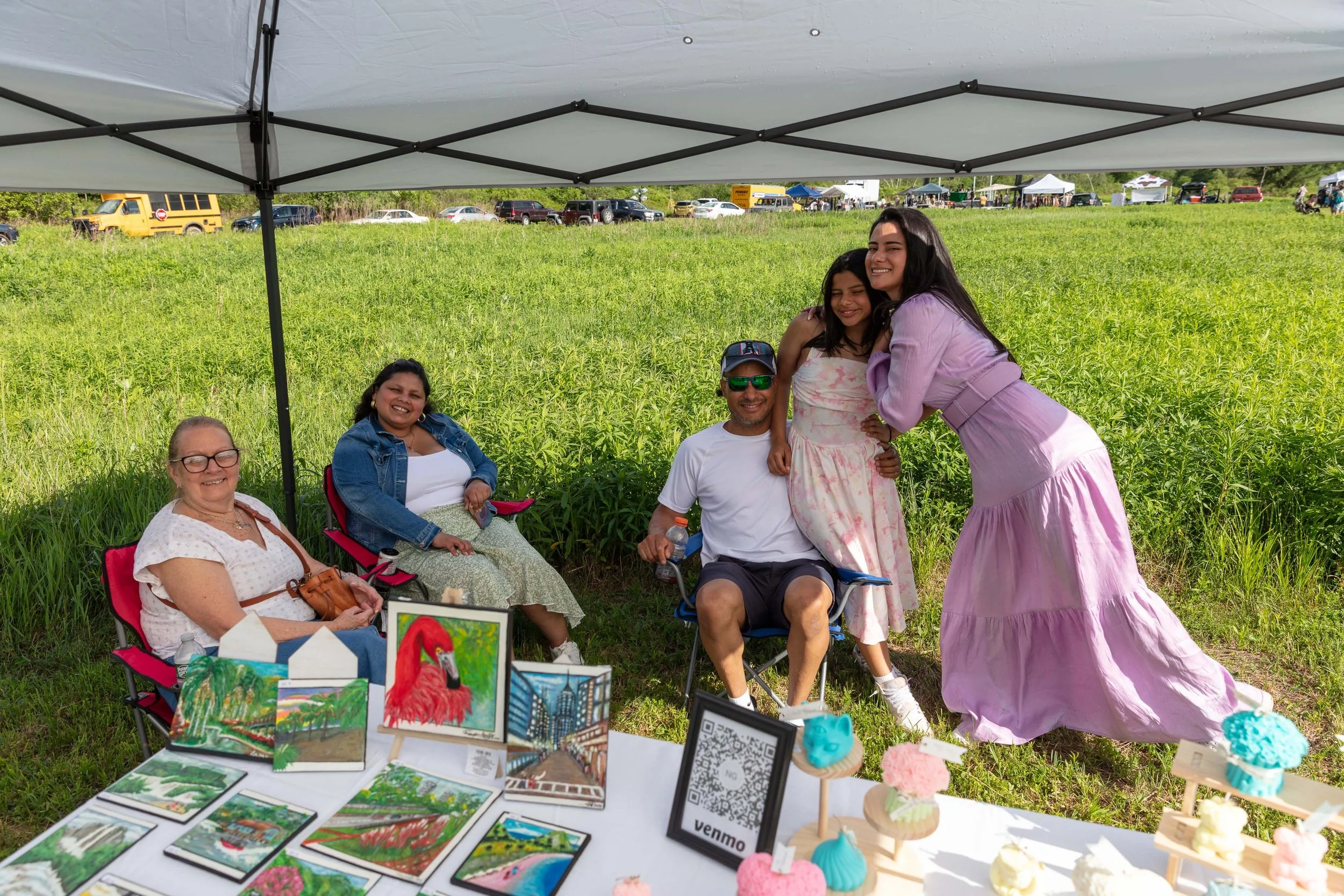 A group of five people, including three women and one man, sitting and standing under a canopy at an outdoor event. They are smiling and enjoying the sunny day with a green field and parked cars in the background. A table with colorful artwork and cr