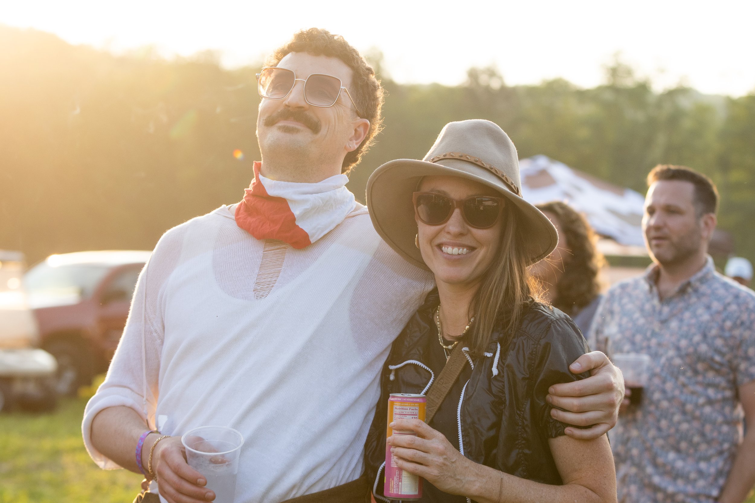 A man and woman at an outdoor event, smiling and hugging, with the sun setting in the background. The man is wearing sunglasses and a white shirt with a red and white bandana around his neck, holding a clear plastic cup. The woman is wearing sunglass