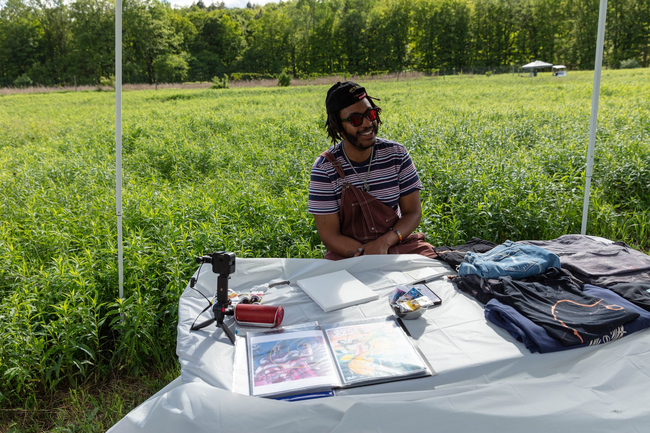 A young man with dreadlocks, wearing sunglasses and a striped shirt, sitting behind a table outdoors in a green field with trees in the background. The table has clothes, a comic book, and art supplies on it, and there is a tent-like cover structure 