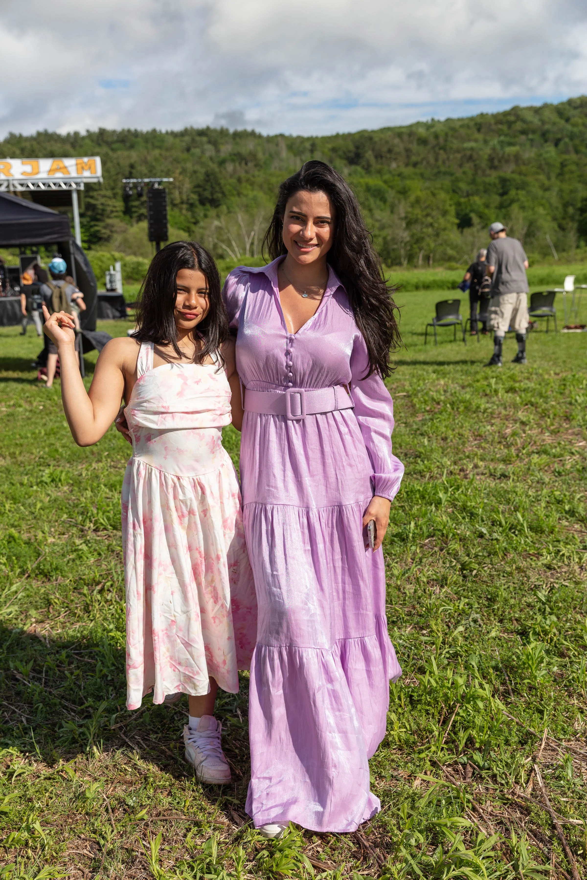 Two young women, one in a pink dress and the other in a lavender dress, standing outdoors on grass with a green hilly background during daytime. People and equipment are visible in the background at an outdoor event.