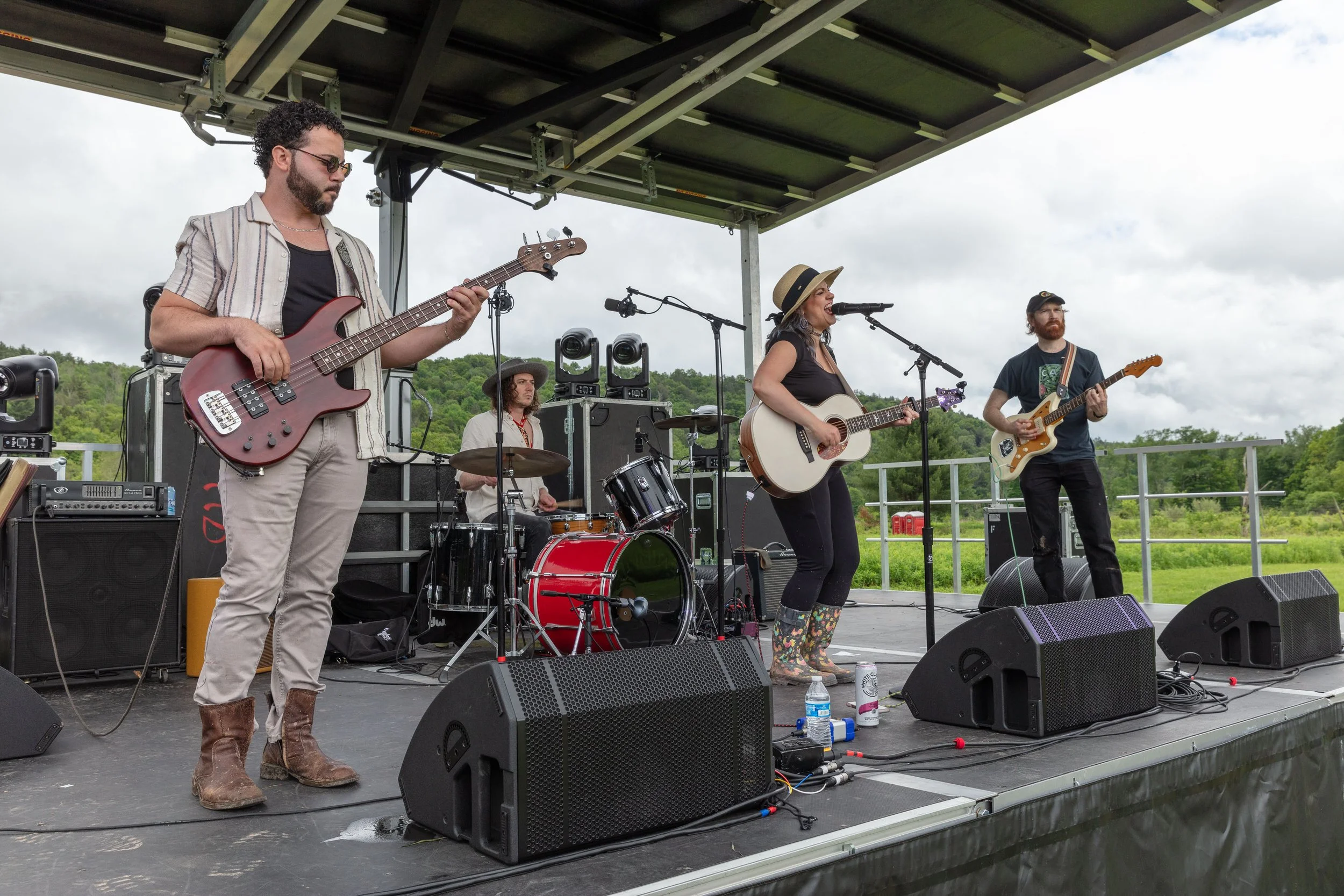 A band performing live on an outdoor stage with four members playing instruments, including guitar, bass, and drums, under a cloudy sky.