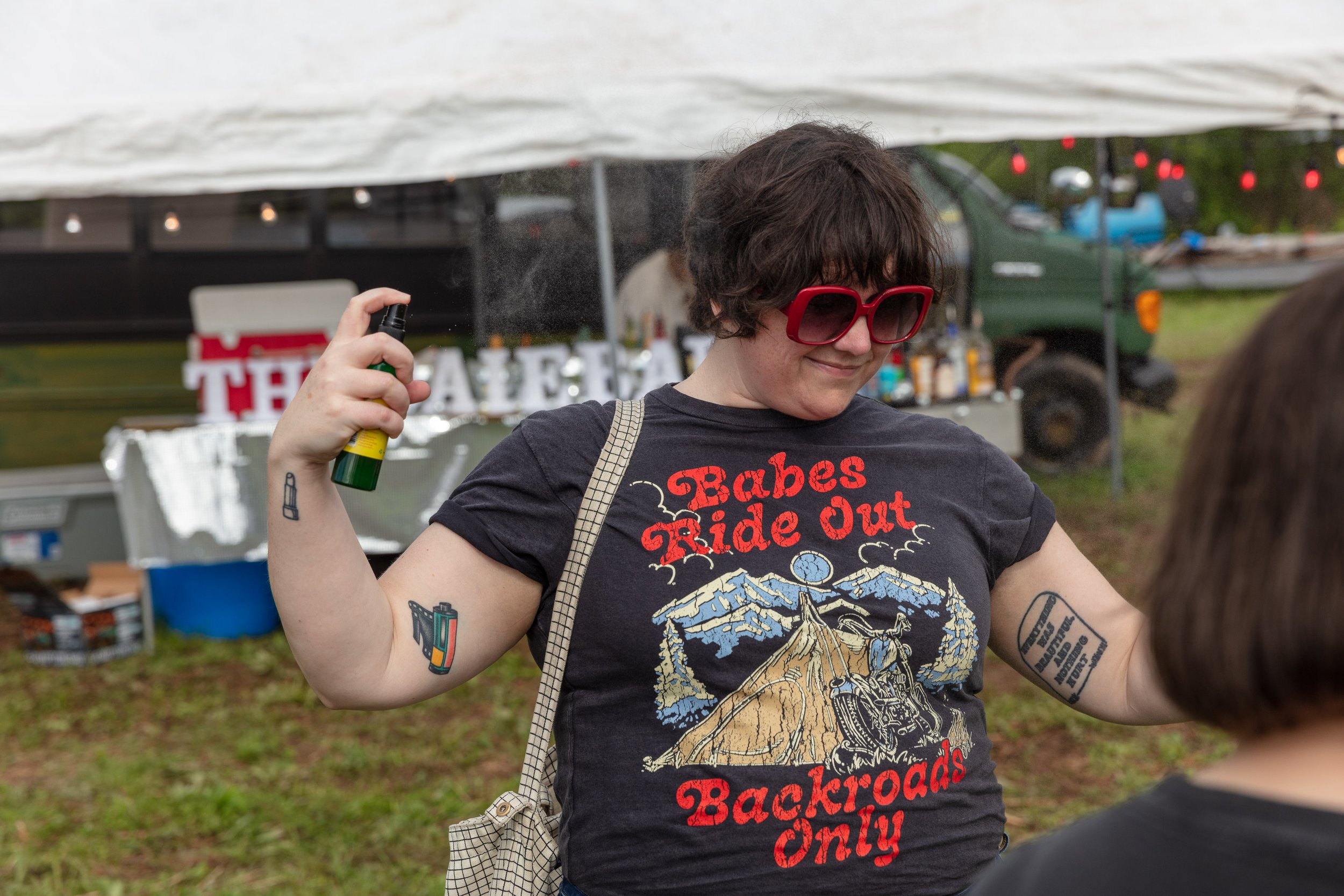 Young woman with curly dark hair wearing red sunglasses and a black T-shirt with a motorcycle illustration and the text 'Babes Ride Out' and 'Backroads Only,' holding a spray bottle in her right hand and smiling outdoors at a gathering or festival.