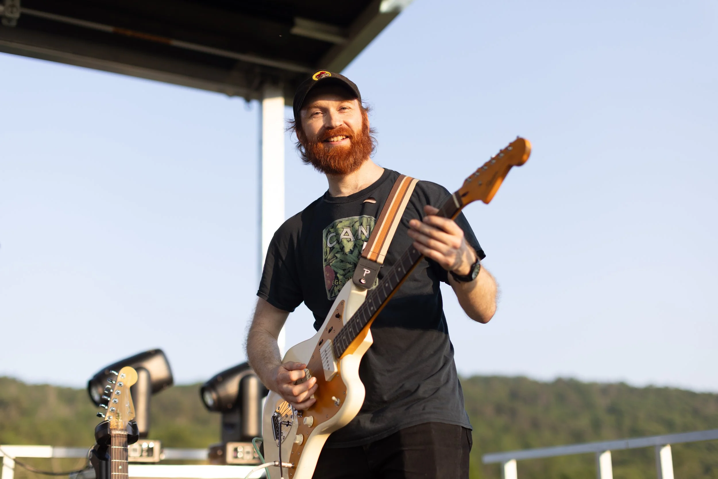 A man with a red beard and mustache wearing a black cap and T-shirt playing an electric guitar outdoors during daytime, with a clear sky and some trees in the background.