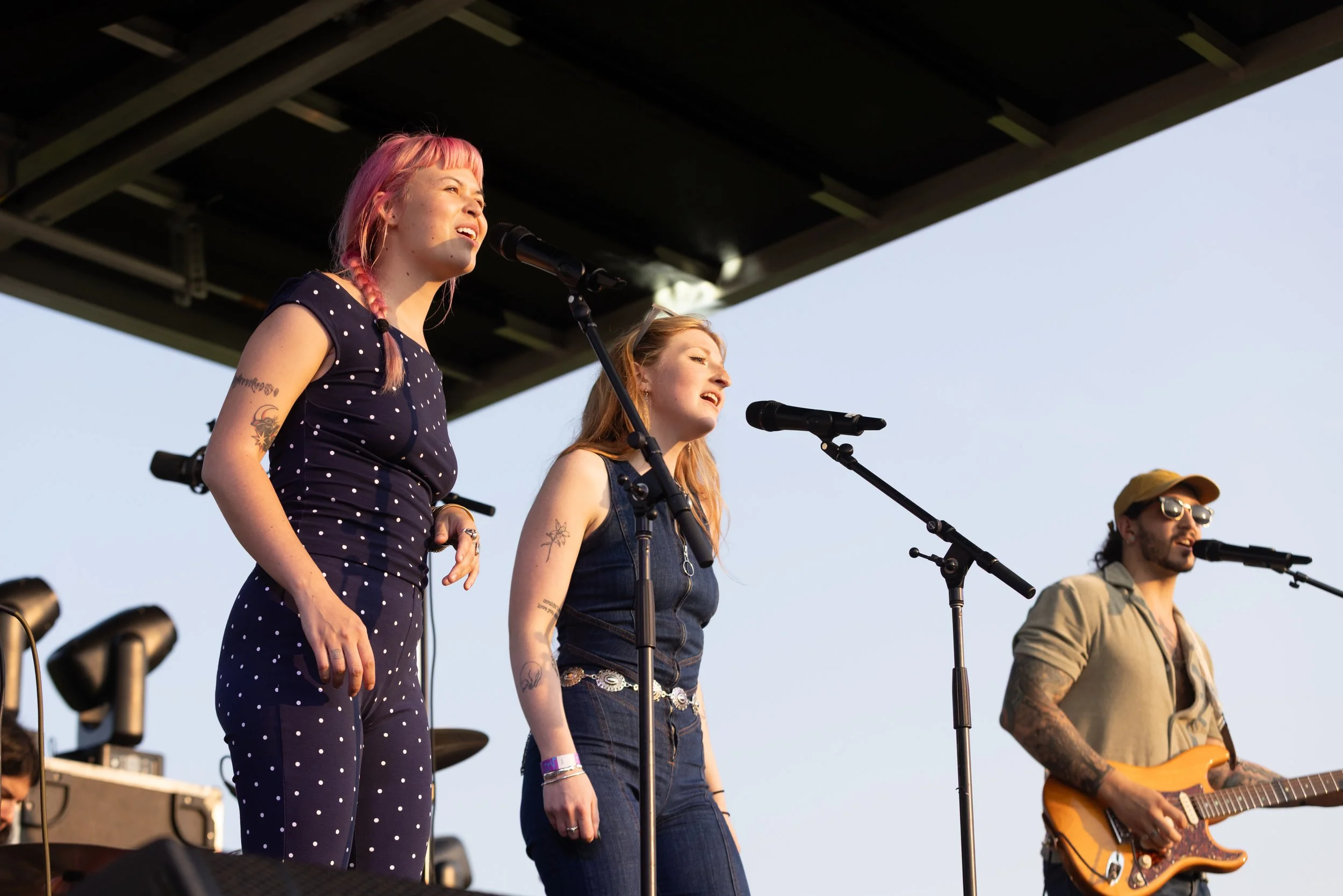 Four musicians perform on stage outdoors. The woman on the left has pink hair, tattoos, and a polka dot dress. Next to her is a woman with red hair, a tattoo on her arm, wearing a denim vest. The third performer is a man with sunglasses and a hat, pl