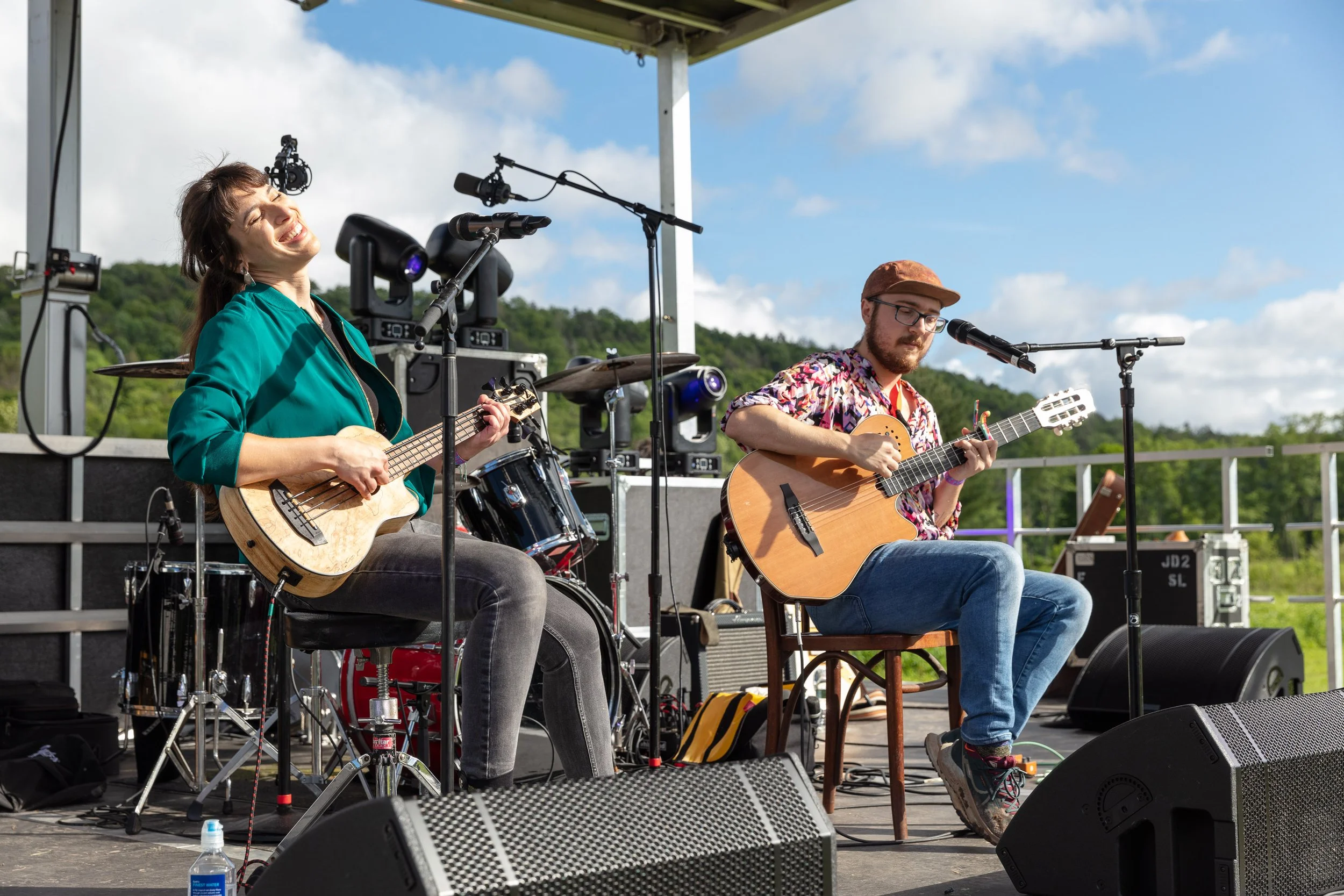 Two musicians performing outdoors on a stage; a woman smiling and playing a ukulele, and a man focused on playing a guitar. There are microphones, speakers, and drums in the background, with a scenic green hill and blue sky behind them.