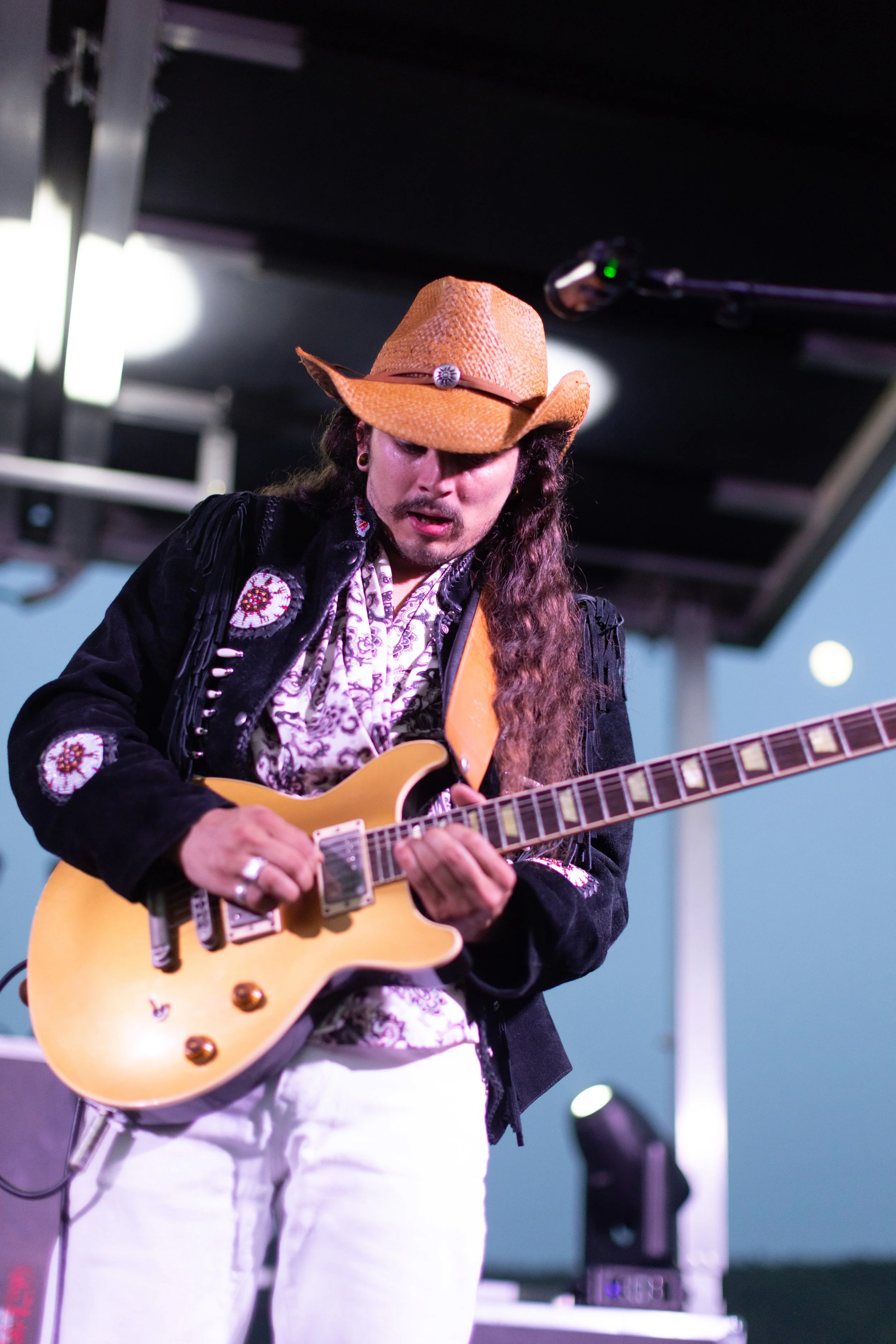 A person playing an electric guitar on stage, wearing a cowboy hat and a black jacket with embroidered patches, white pants, and a patterned shirt.