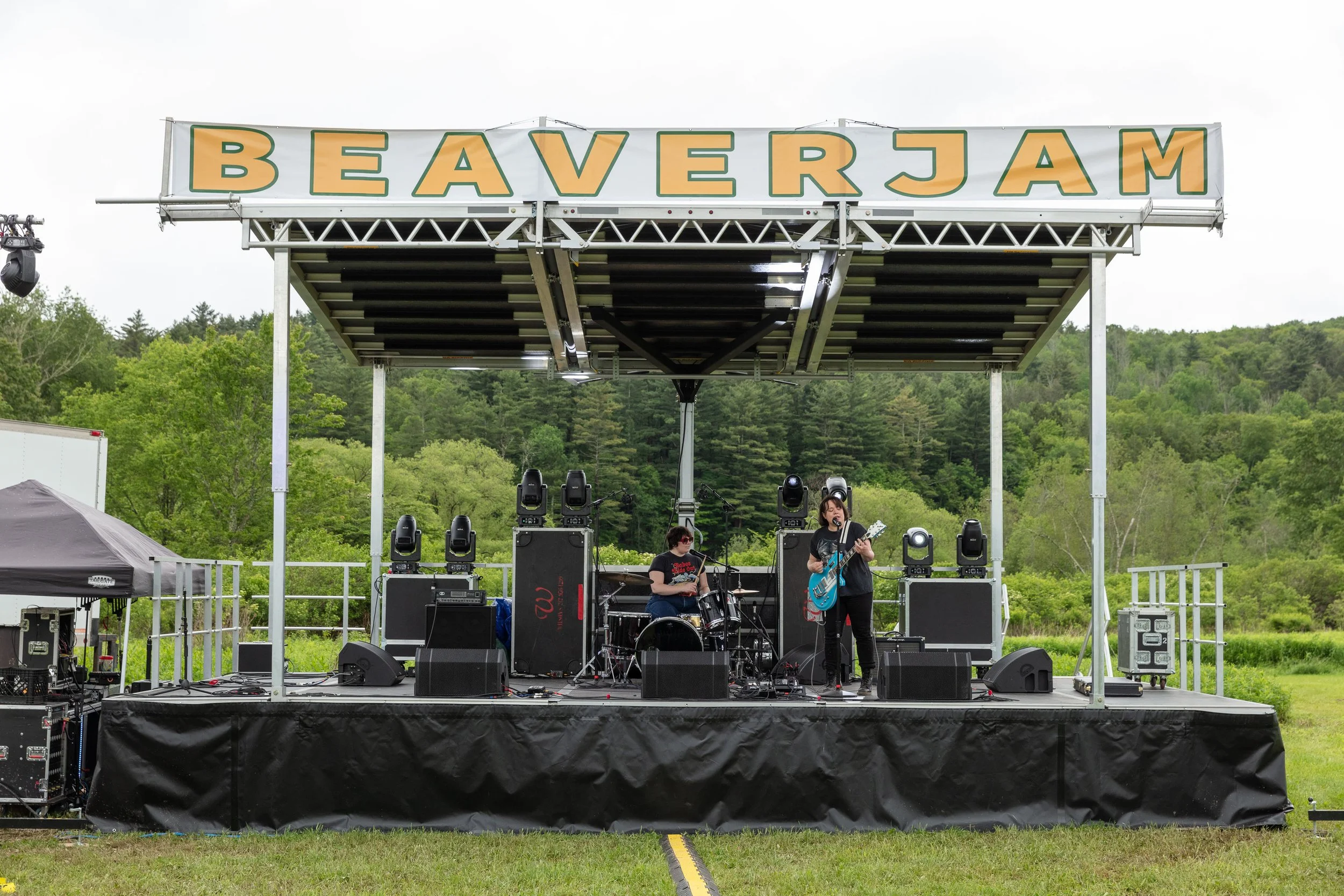 An outdoor stage with a band performing at a festival called Beaver Jam, with trees and hills in the background.