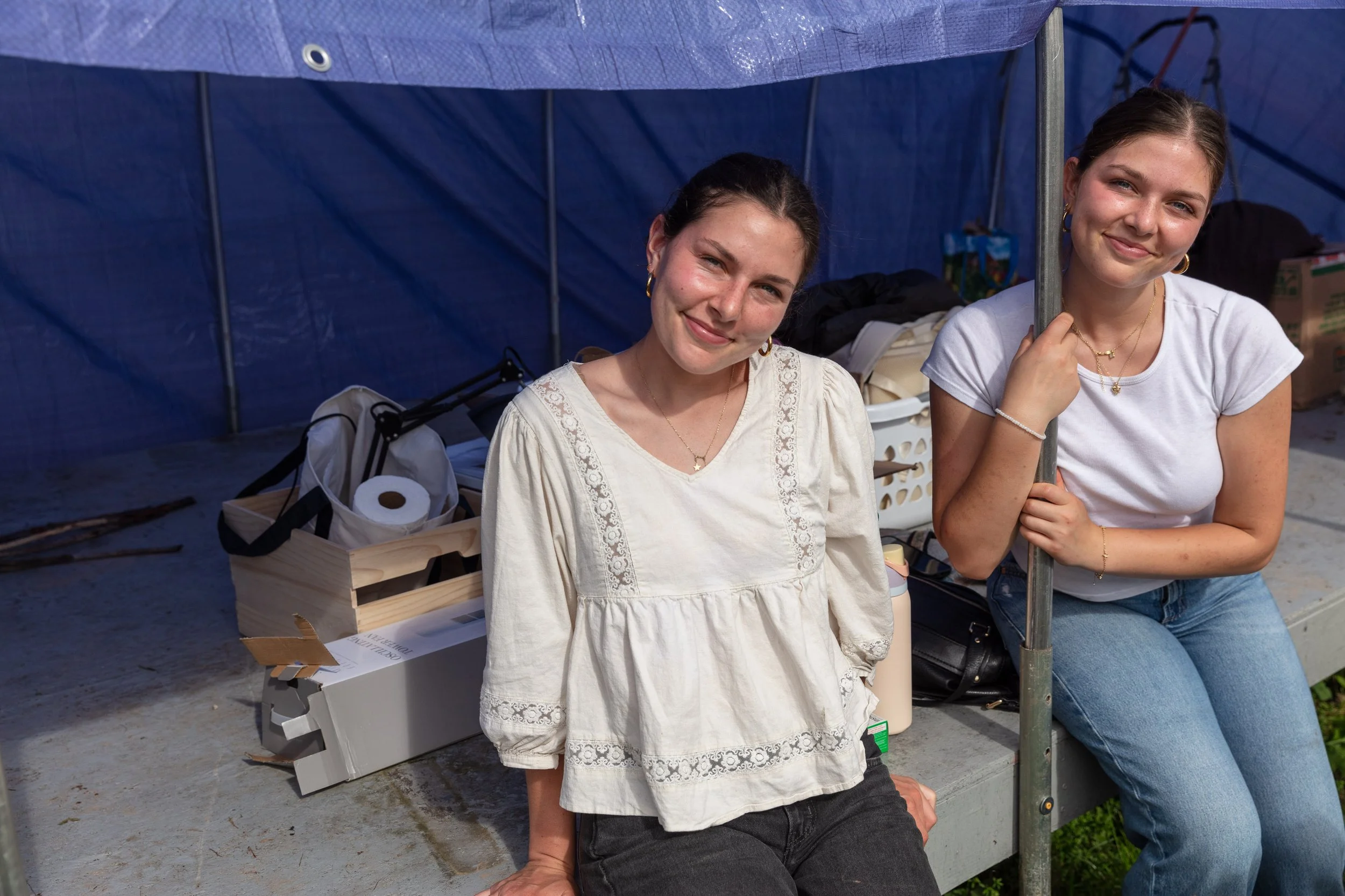 Two young women sitting outdoors under a blue canopy, smiling at the camera, with various items and boxes around them.