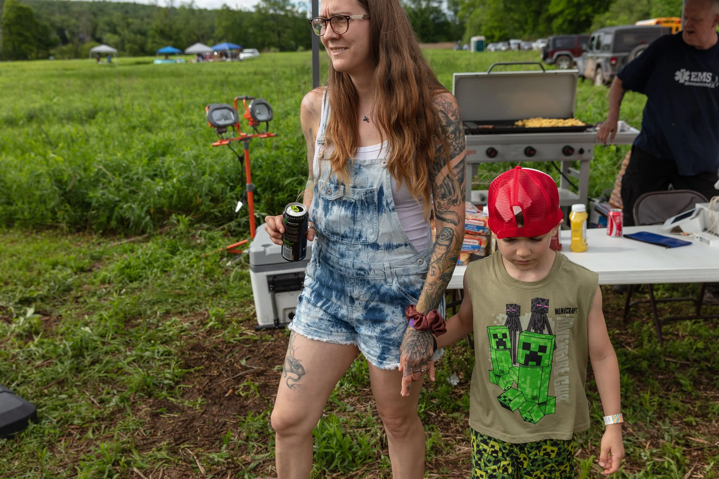 A woman with long hair, glasses, and tattoos, holding a soda can, walking hand-in-hand with a young boy wearing a Minecraft shirt and patterned shorts, during an outdoor event at a grassy park with tents and vehicles in the background.