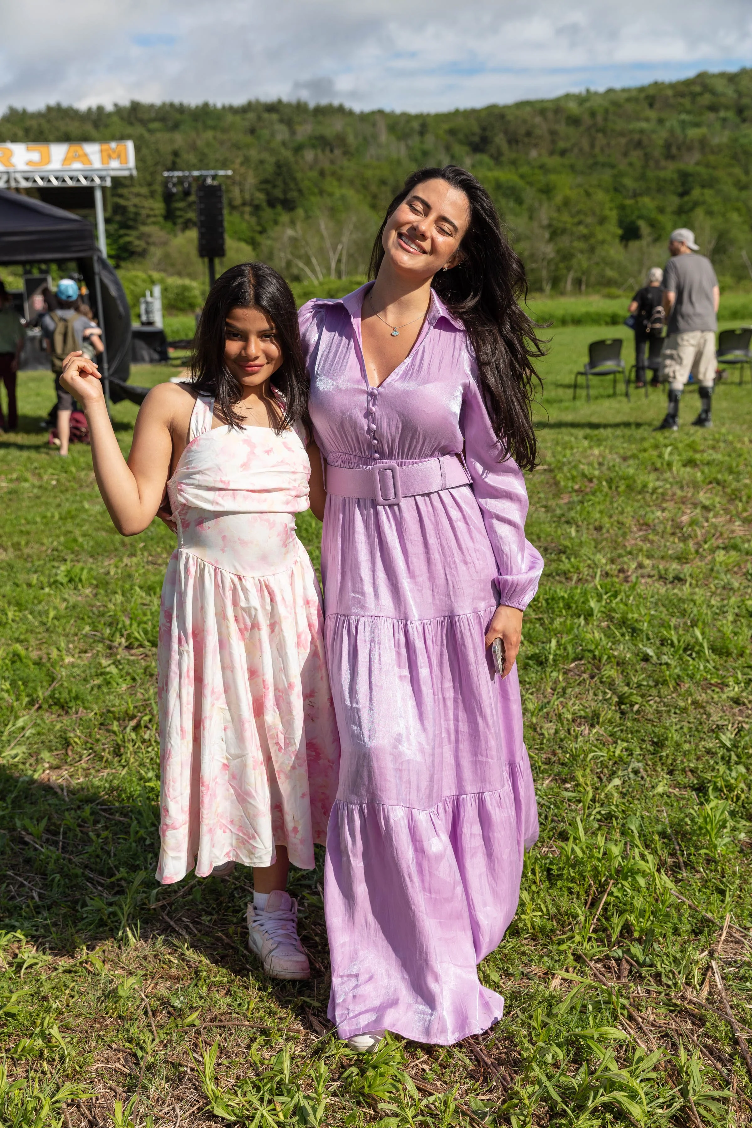 A woman and a girl smiling outdoors on a grassy field with a stage and people in the background. The woman wears a light purple dress, and the girl wears a light pink dress with a floral pattern.