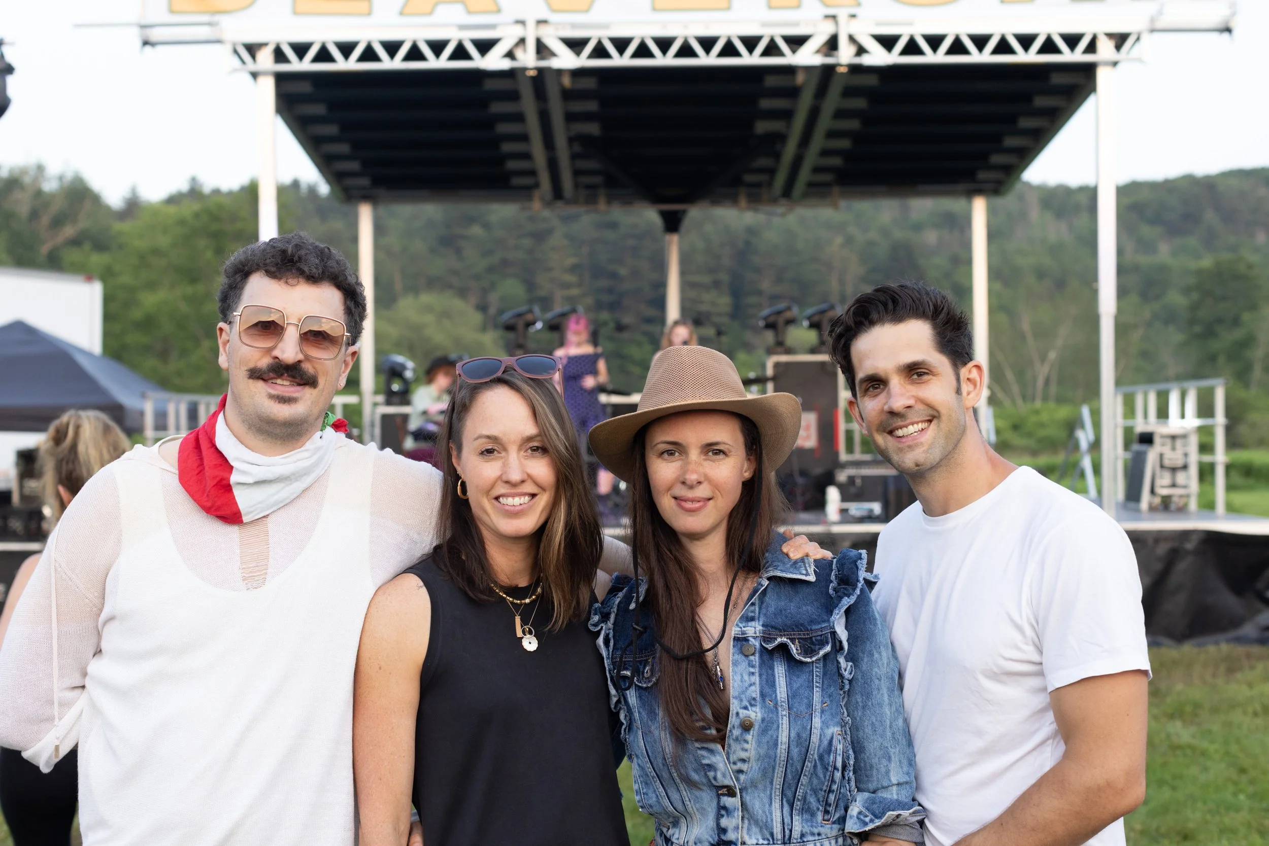 Group of five people posing outdoors at a music festival with a stage and green trees in the background.