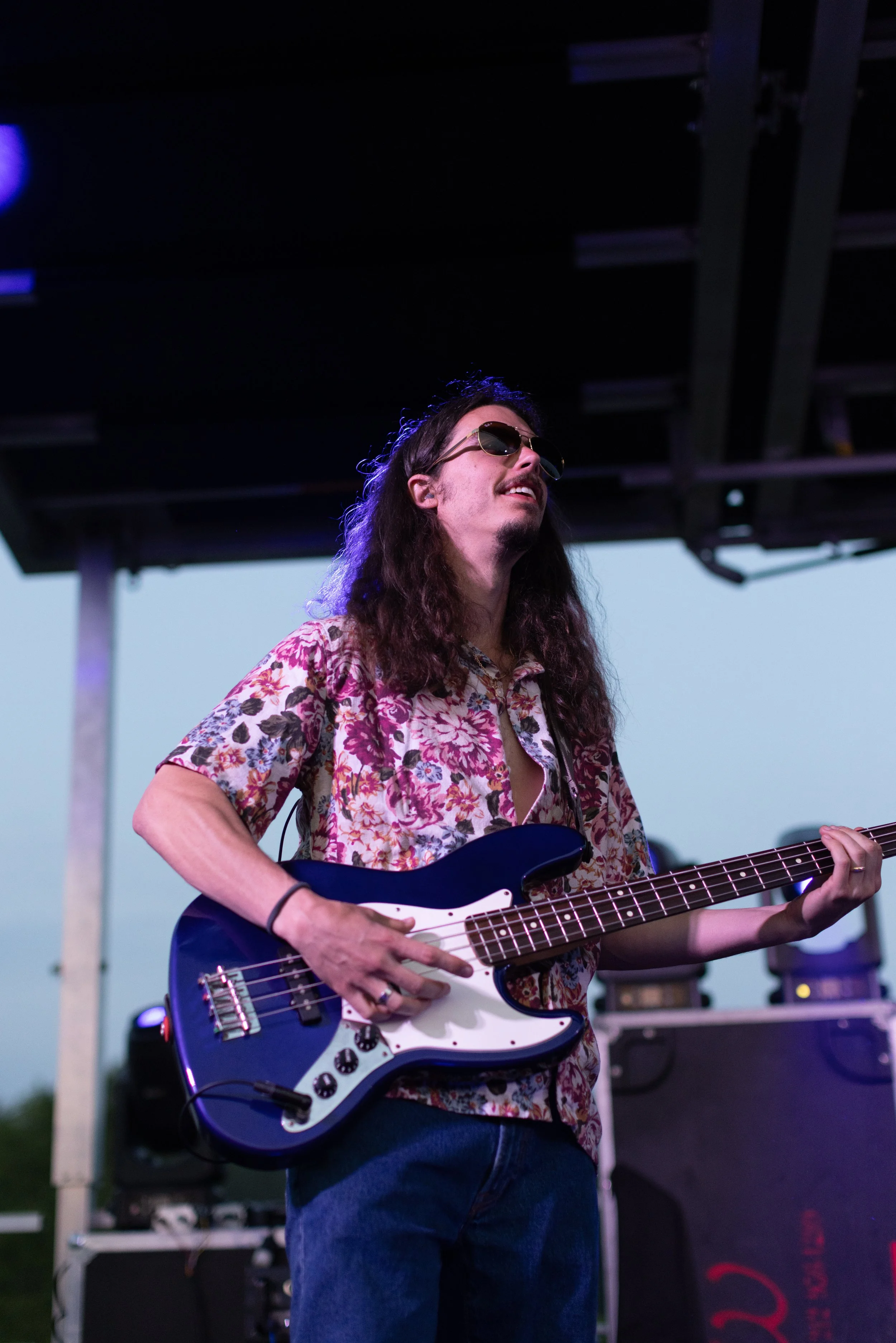 Person playing electric guitar on stage at outdoor event, wearing sunglasses and floral shirt, dark background.